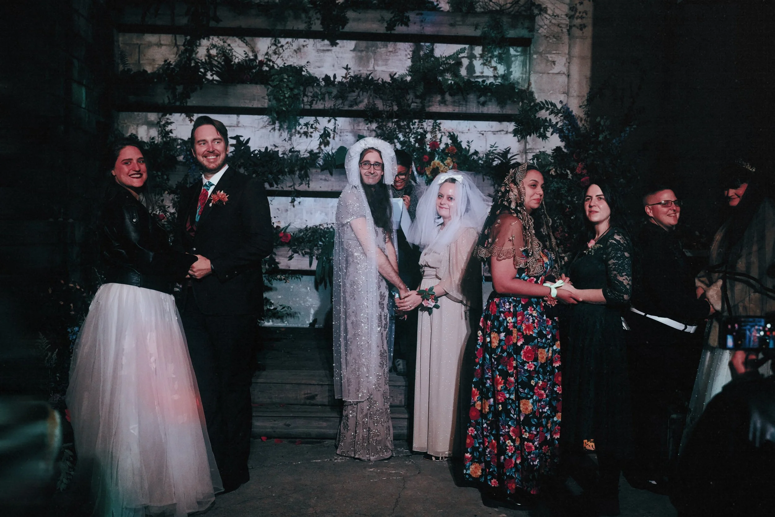 A group of people at a wedding, with some in wedding attire and others in diverse outfits, standing in front of a rustic wooden wall with greenery and flowers, holding hands and smiling.