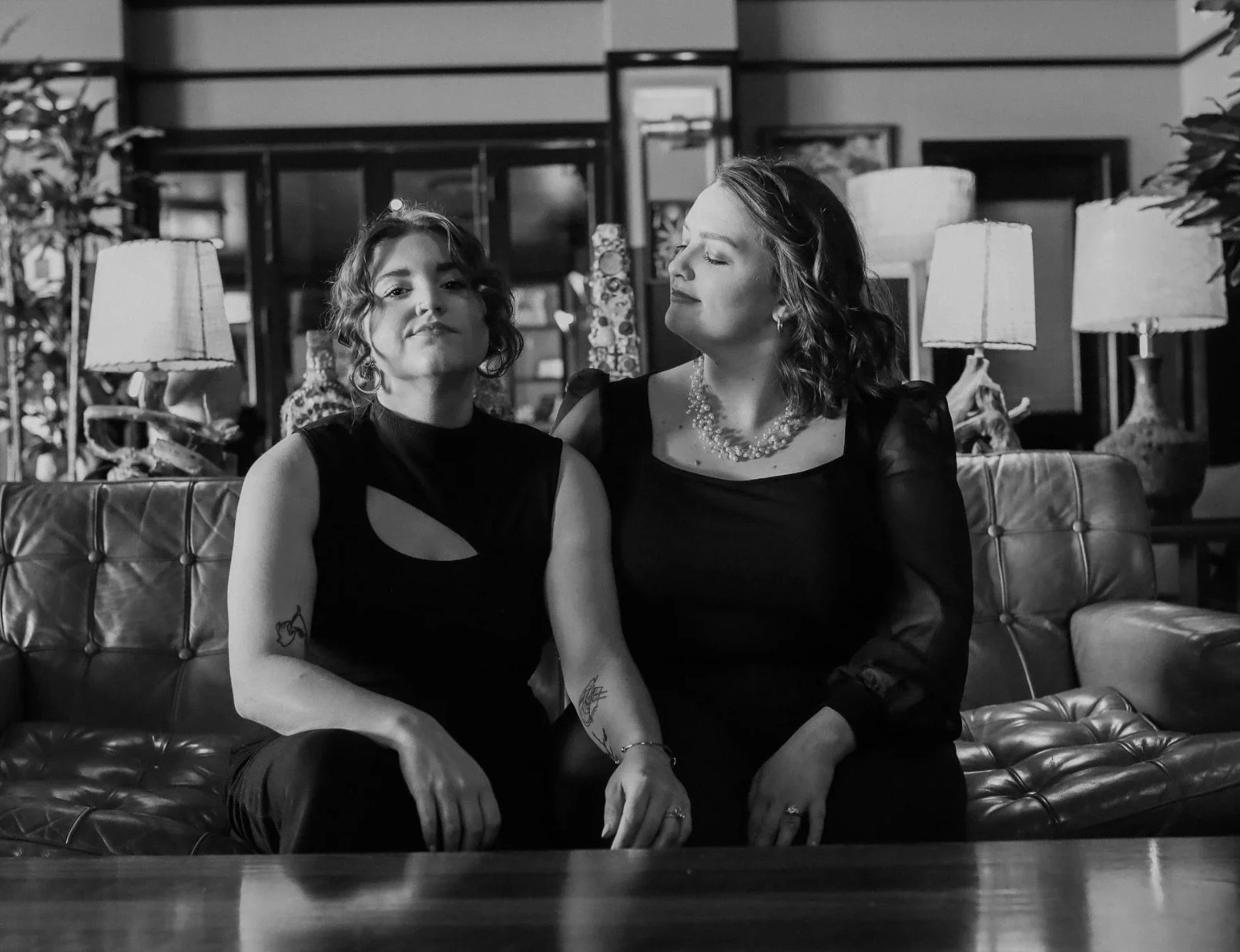 Two women sitting on a leather couch in a stylish room with lamps and artwork, one looking directly at the camera with a confident expression, the other with a relaxed, closed-eye expression.