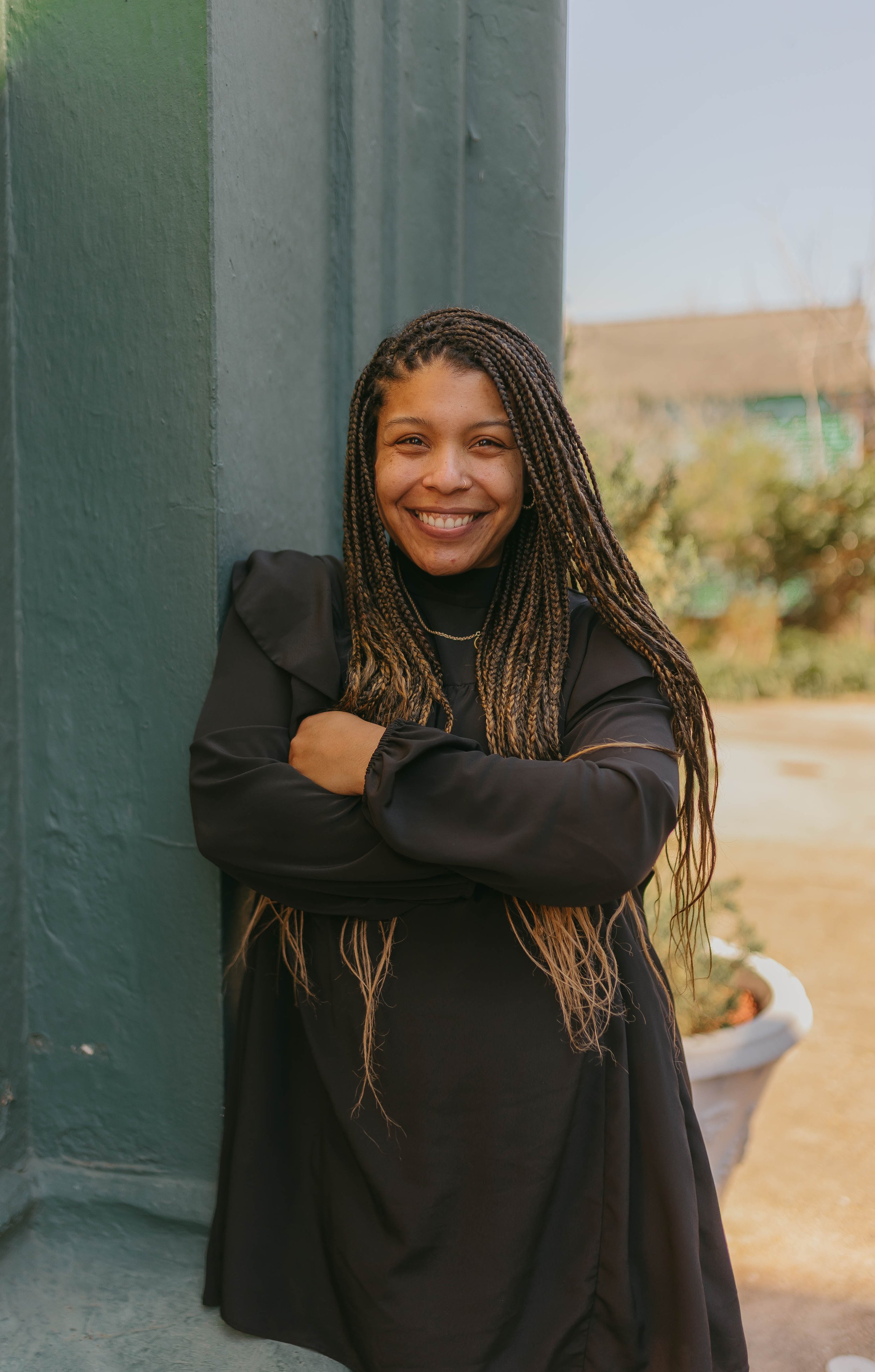 A young woman with braided hair, smiling, standing outdoors leaning against a green wall with arms crossed.