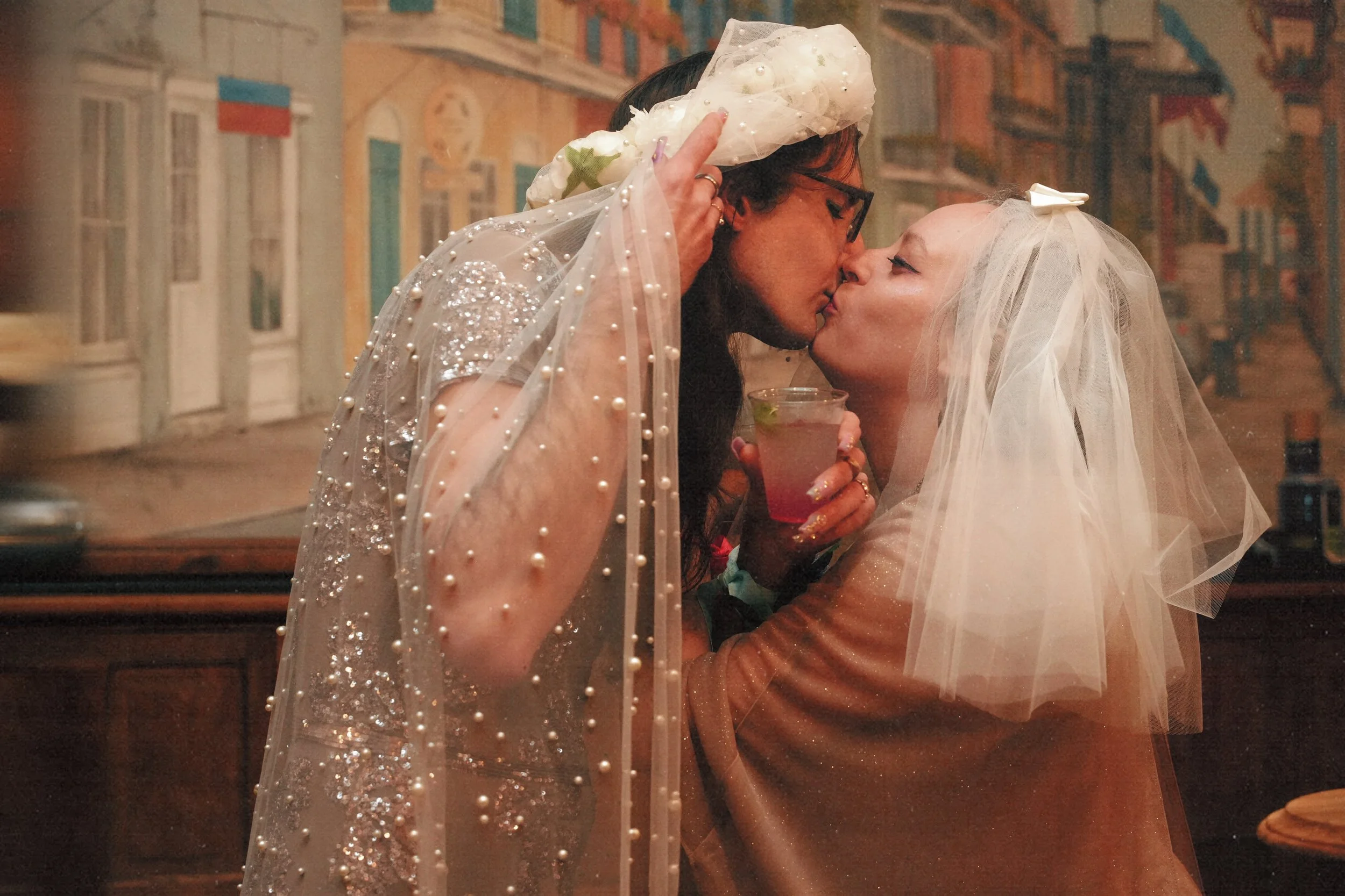 Two women, dressed as a bride and groom, are sharing a kiss at a reception. The woman on the left is wearing glasses and a dress with pearl details, while the woman on the right is dressed in a wedding veil and holding a drink.