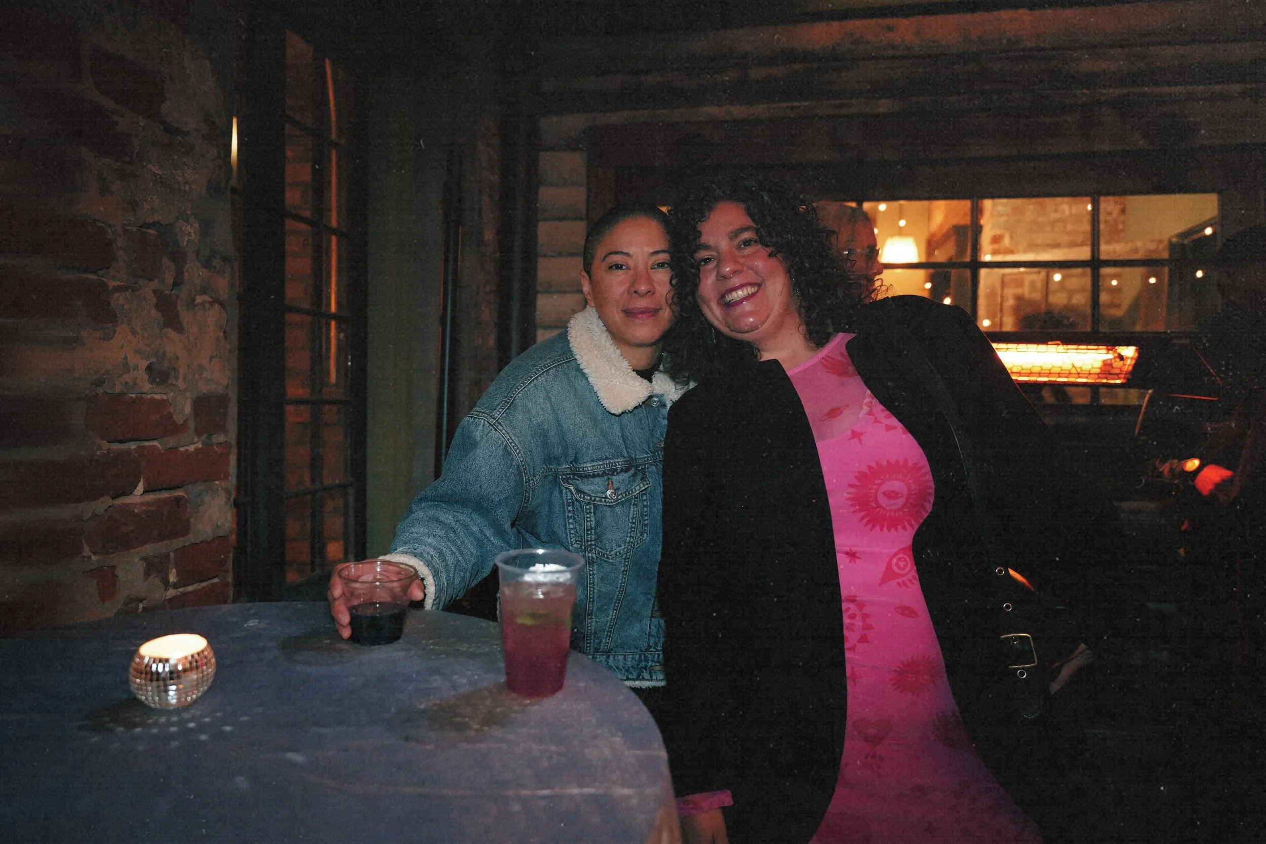Two women smiling and posing for a photo at a bar or restaurant, with drinks on the table and warm lighting in the background.