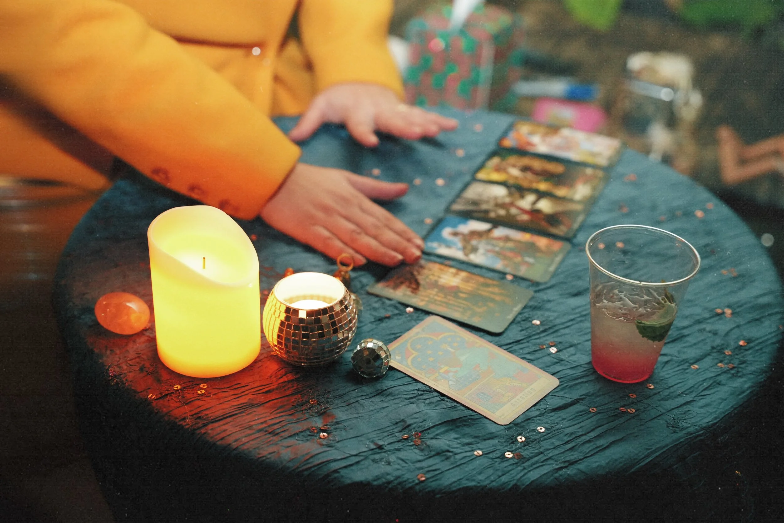 A tarot card reading setup with a person in a yellow jacket reading Tarot cards on a dark blue table. The table is decorated with small shiny embellishments, a lit yellow candle, circular and disco ball candles, and a glass of pink drink with mint.