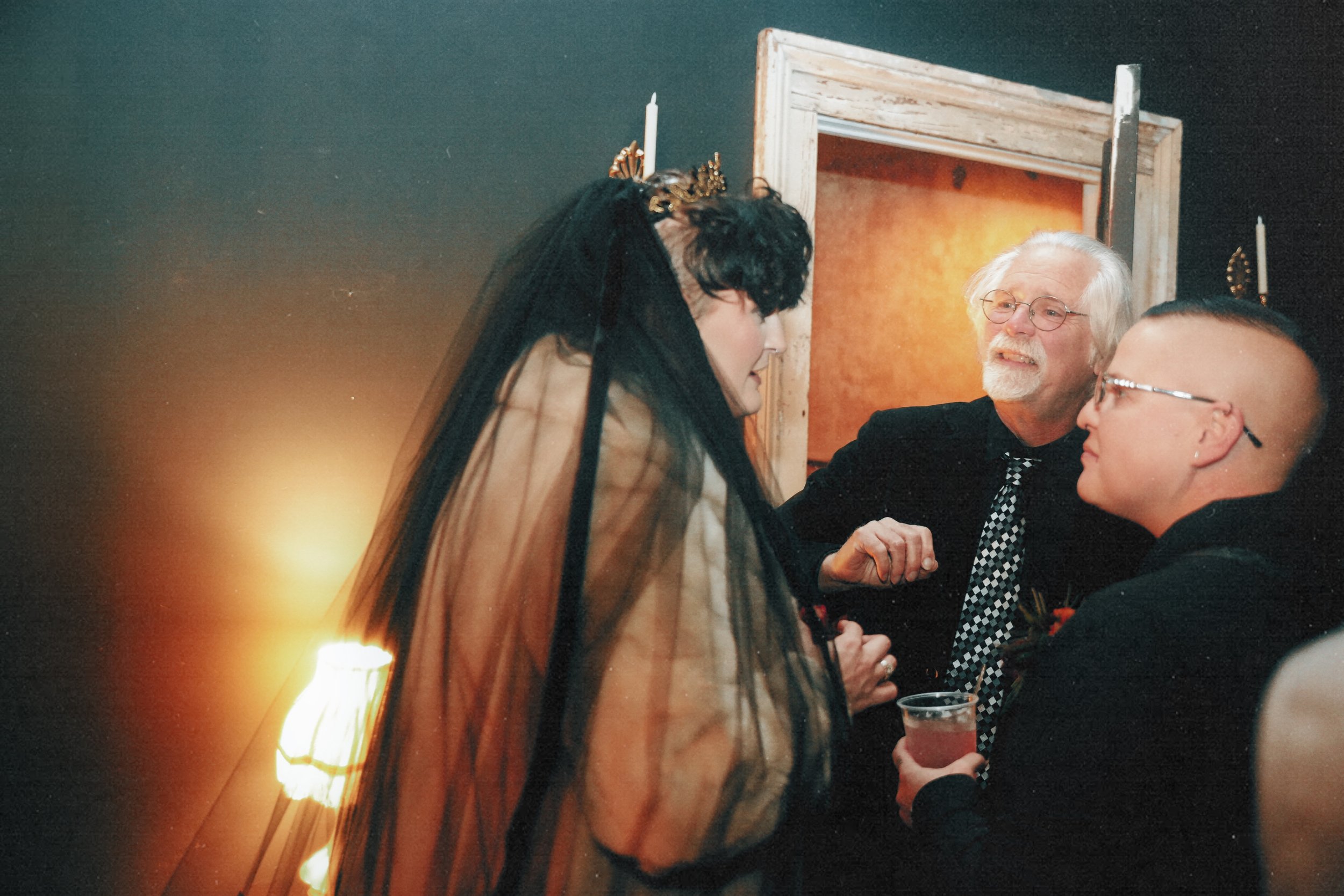 Three people at a party, two men and a woman, are engaged in conversation with the woman dressed in a black veil and the man with white hair and beard smiling. The man with a shaved head and glasses is holding a drink.