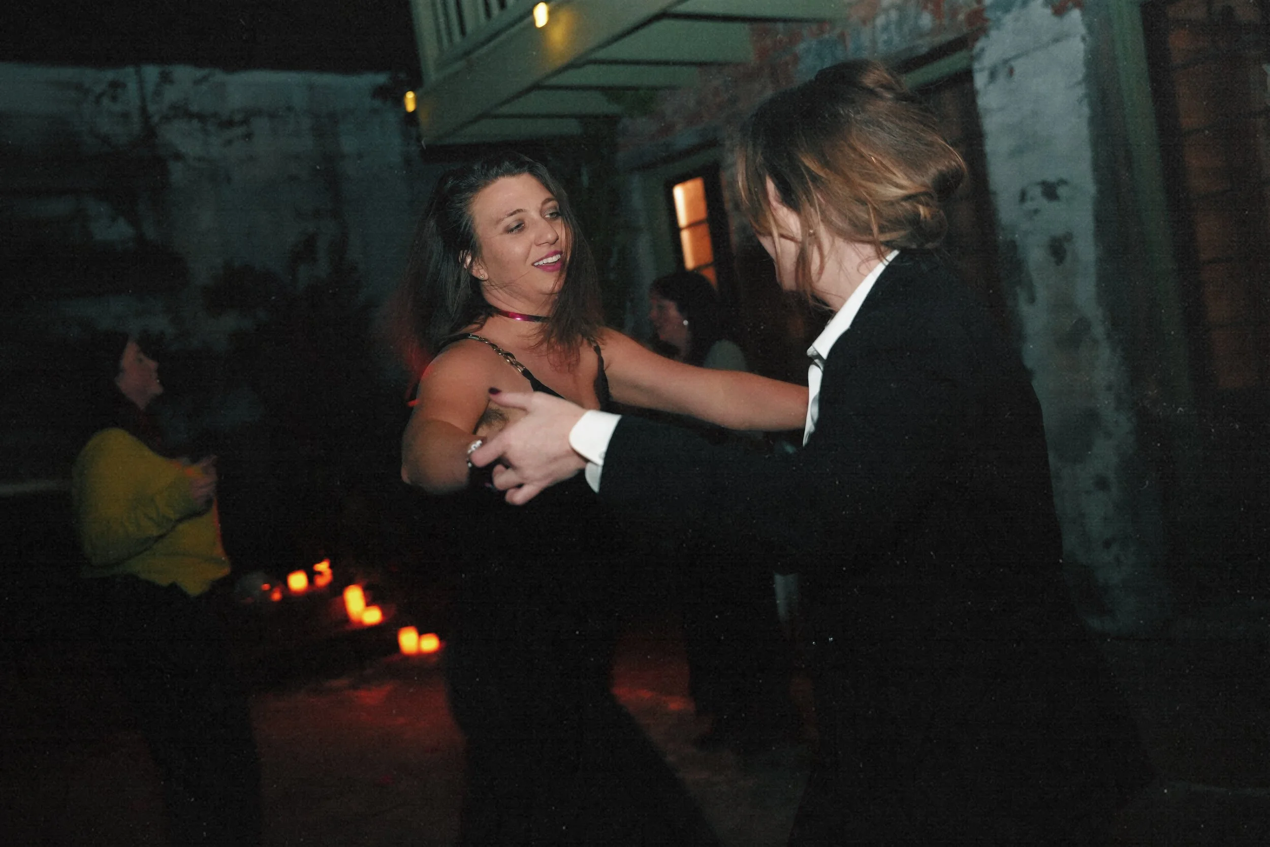 A woman and a man are dancing together at a party or nightclub, with the woman smiling and the man facing away from the camera. There are other people in the background and candles on a table, creating a dimly lit ambiance.
