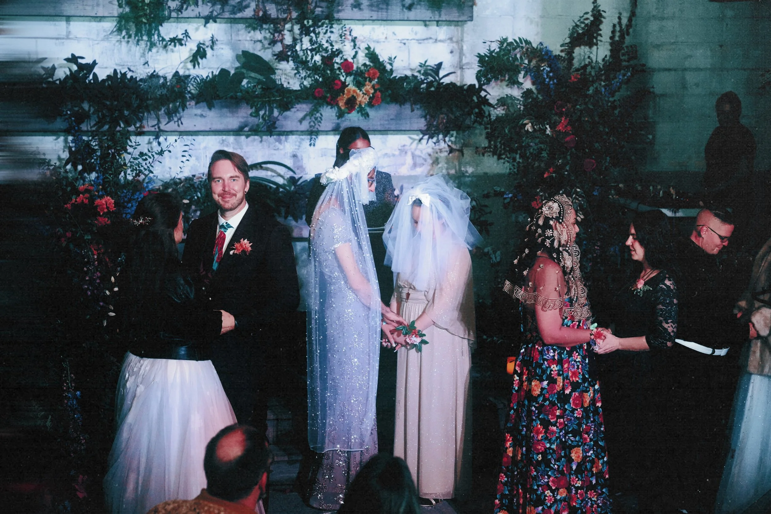 A wedding ceremony with two brides and two grooms standing in front of a decorated backdrop with flowers, holding hands, surrounded by guests, in an indoor setting.