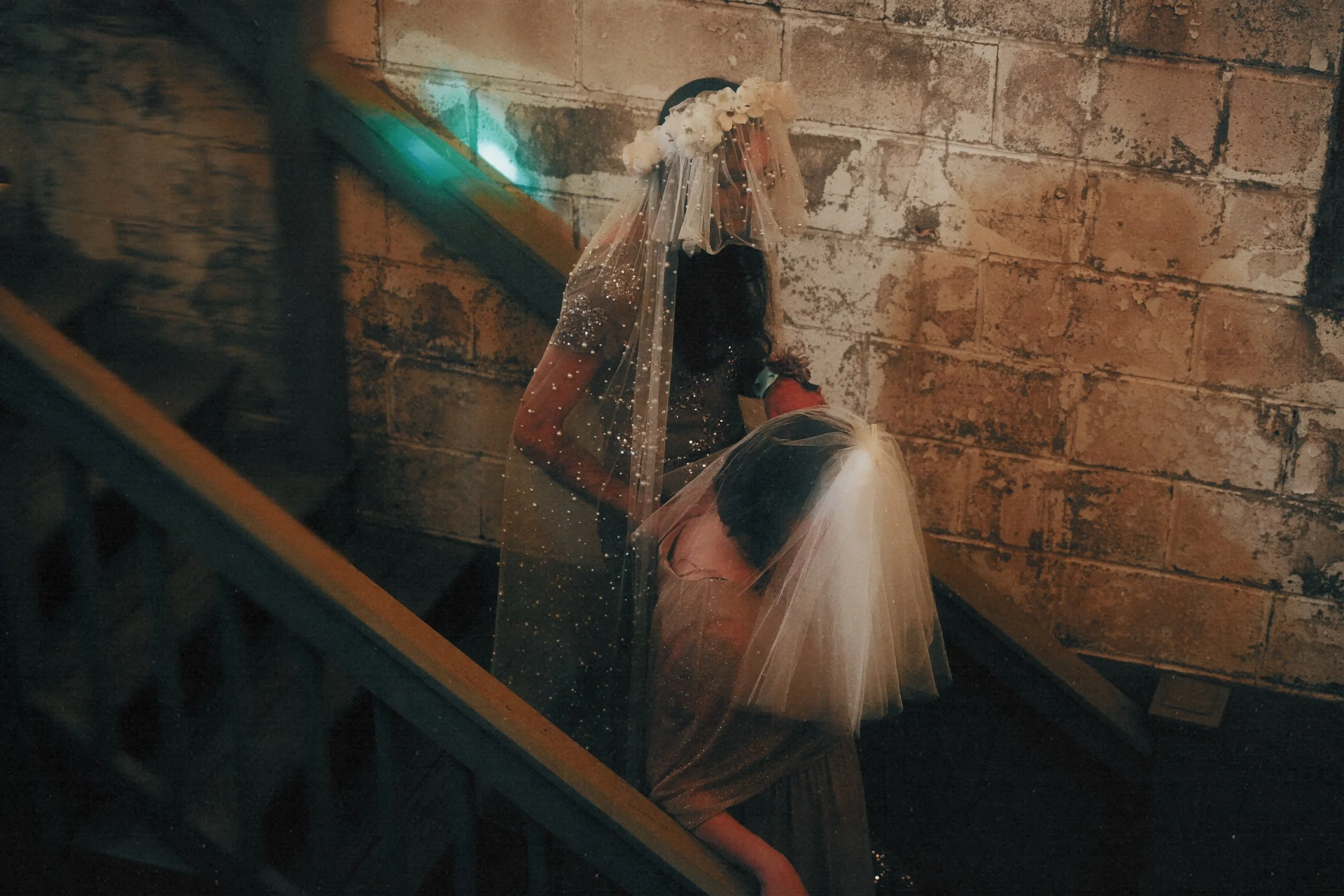 Two women dressed as brides descending a stone staircase, wearing white tulle veils, with one holding the other's arm, in a dimly lit setting with brick walls in the background.