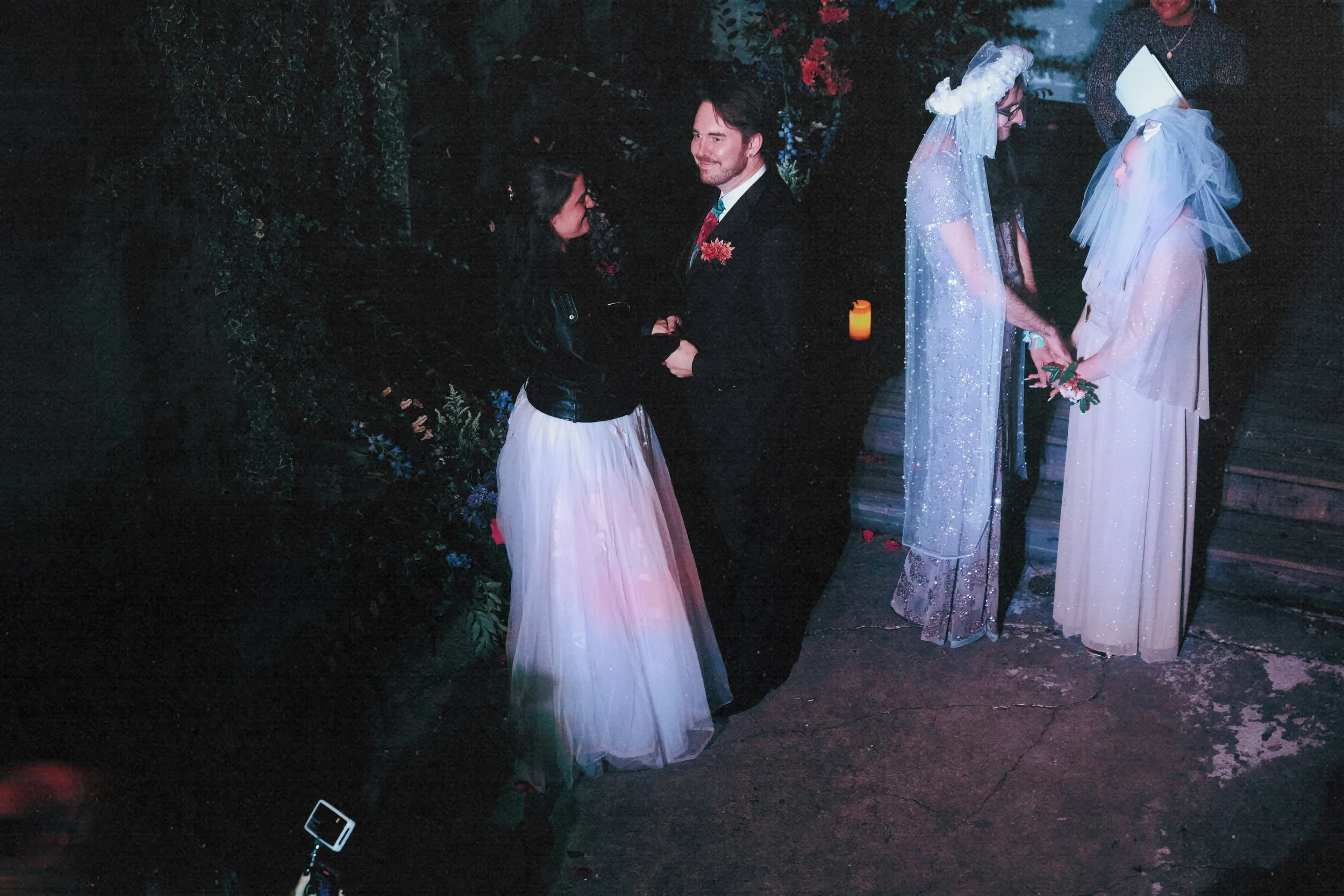A wedding ceremony with two couples. The couple on the left is standing outside under dark trees, holding hands and looking at each other. The couple on the right is standing under a light, with women wearing veils and holding flowers. The ground is 