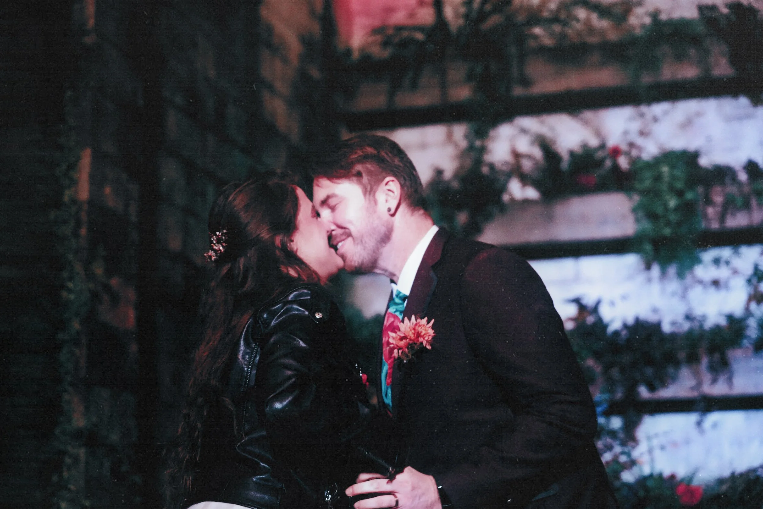 A couple sharing a kiss in front of a brick wall with greenery, the woman wearing a black leather jacket and the man dressed in a suit with a colorful boutonniere.