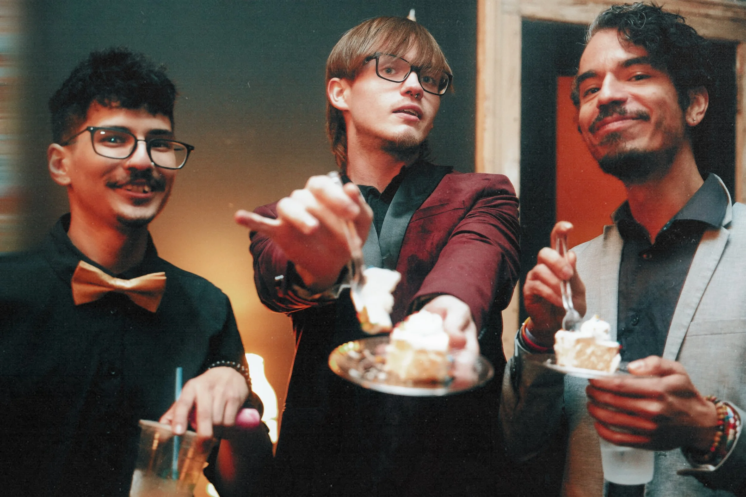 Three men at a party, one with a bow tie and glasses, one with long hair and glasses, and the third with a dark curly hairstyle, all holding plates or glasses of cake, smiling and enjoying the celebration.