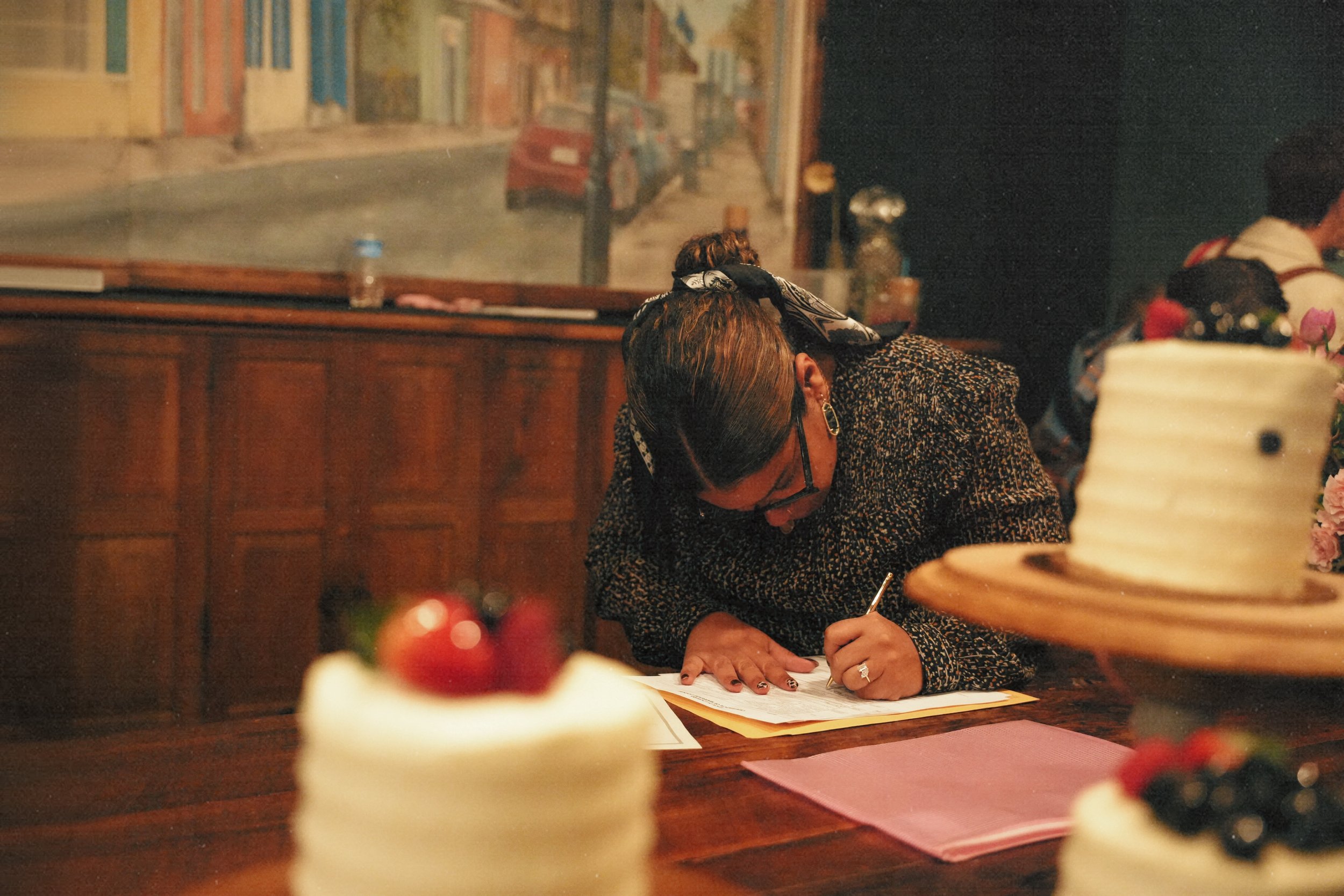 A person with short hair, glasses, and earrings is leaning over a table, writing on a piece of paper with a pen. The person is wearing a dark, patterned top and a scarf. In the foreground, there are cakes with fruit toppings. The background shows a m