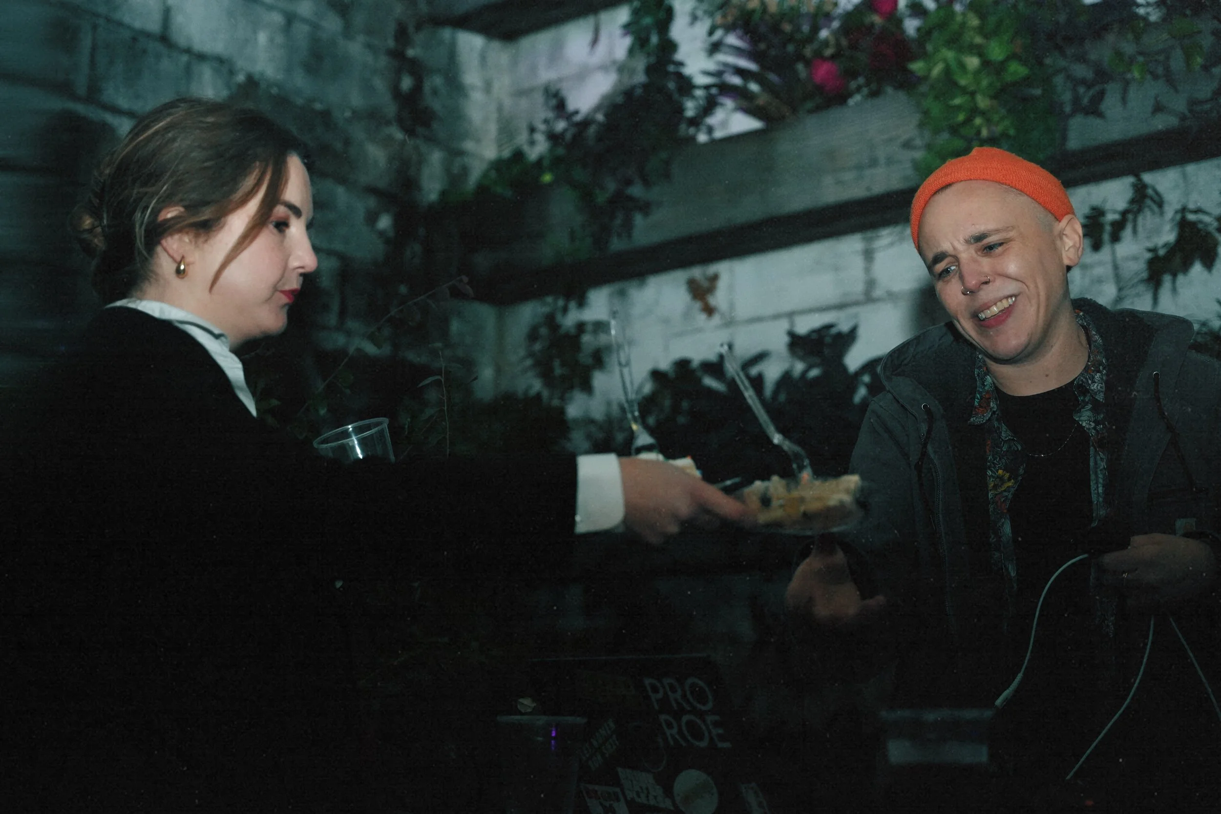 A person with a shaved head wearing an orange beanie is smiling and receiving a slice of pizza from a woman at night outdoors. The woman is dressed in dark clothing and holds a glass.