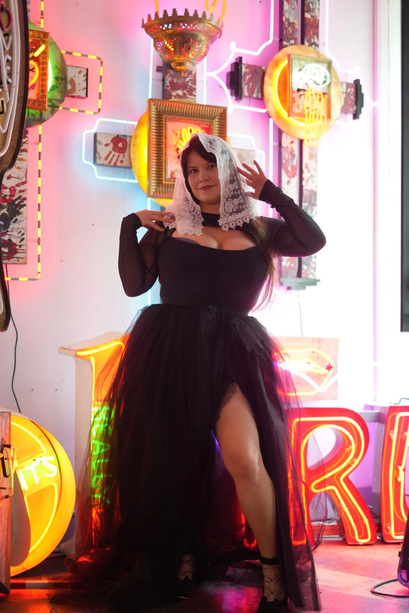 Woman wearing a black dress with a tulle skirt and lace veil posing in front of neon signs and artwork.