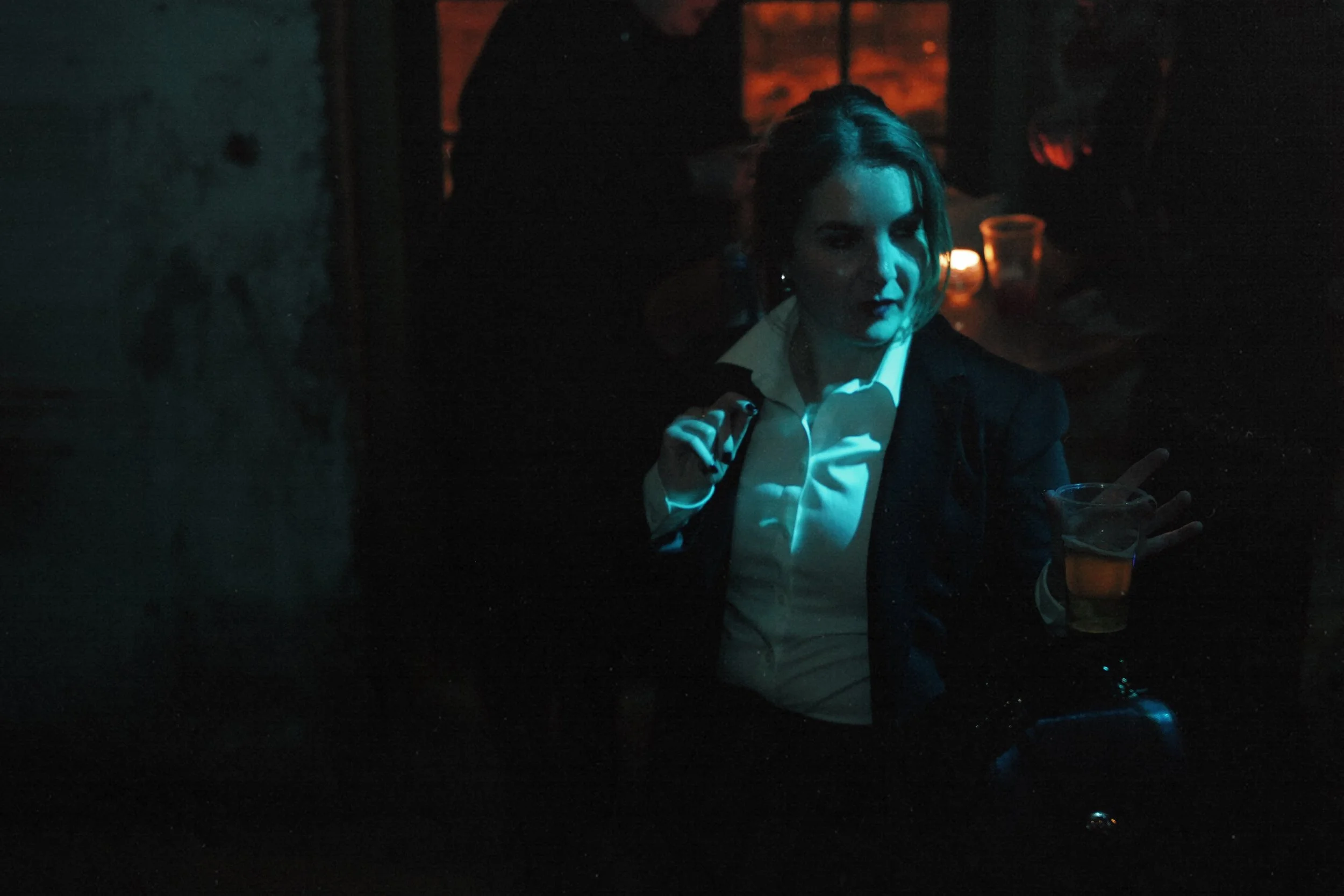 A woman in a white shirt and dark jacket holding a drink, standing in a dimly lit space with candles, looking towards the camera.