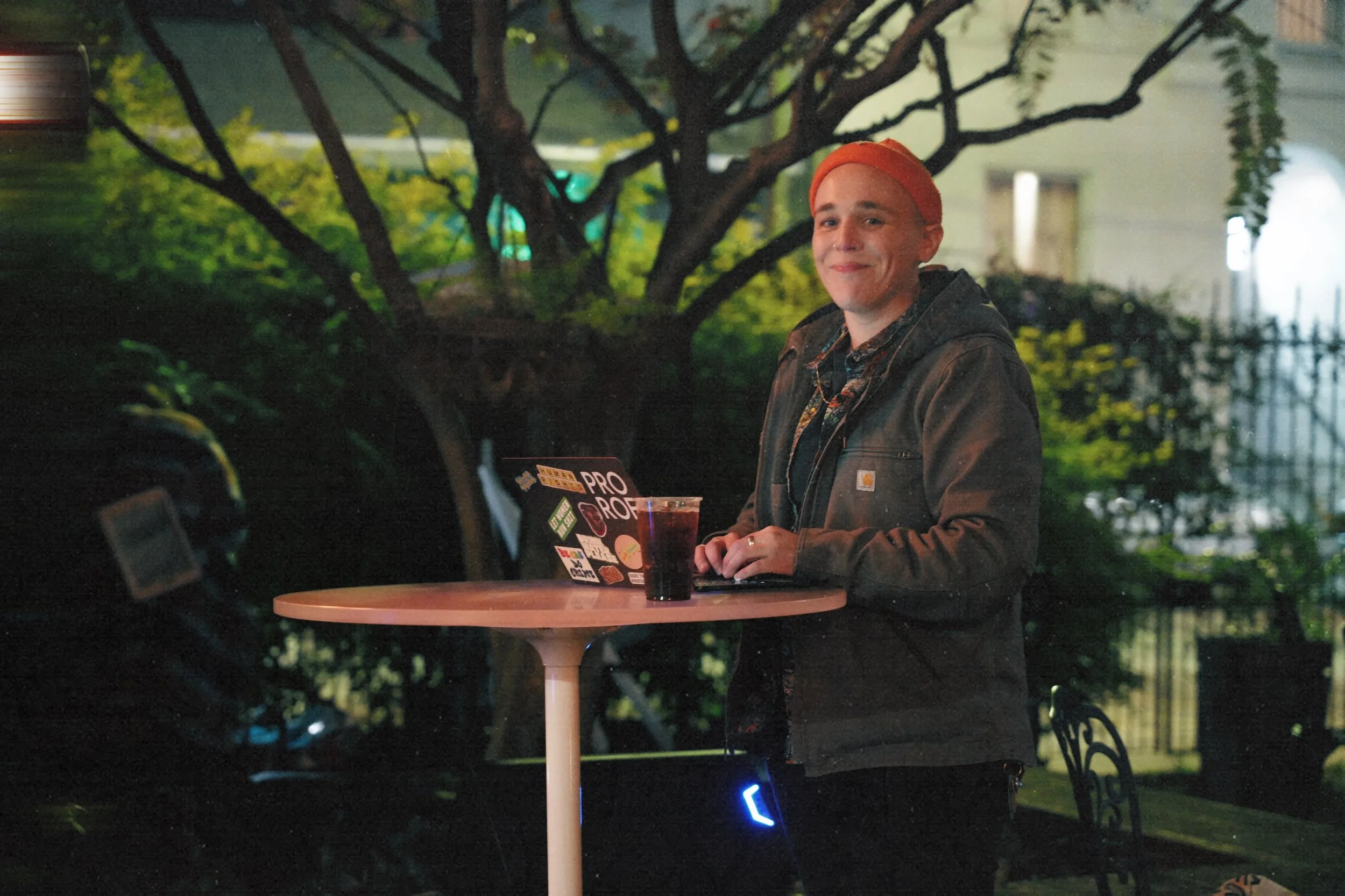 A person standing outdoors at night, smiling, wearing a red beanie and a dark jacket, standing at a round table with a laptop covered in stickers and a glass of dark beverage. There are trees and a building in the background.