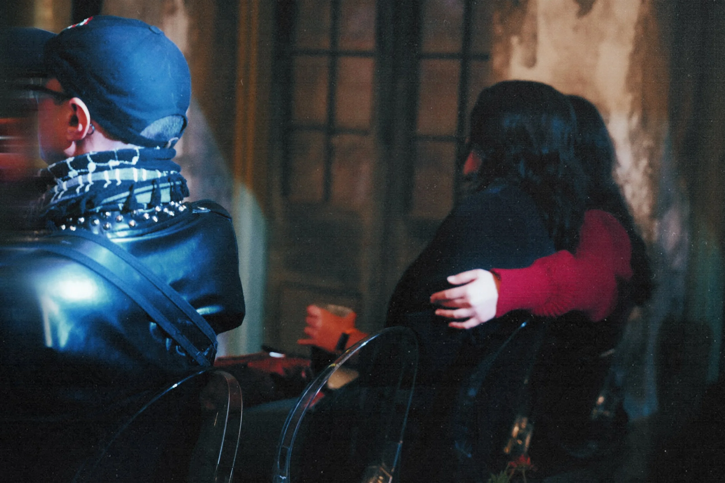 Three people sitting on chairs in a dimly lit room with stone walls and a window. Two are visible from the back, one with a black jacket and red sleeves, the other with a leather jacket and a checkered scarf. The third person's face is not visible.