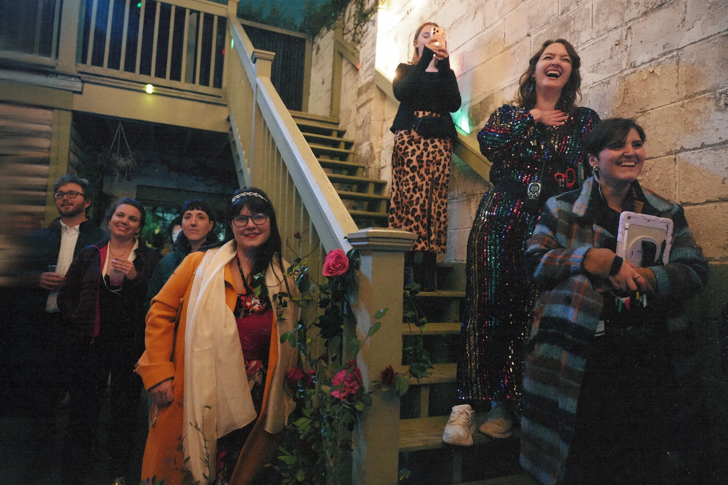 A group of seven people are gathered on and near a staircase, laughing and taking photos at a social event or party in a warmly lit indoor setting.