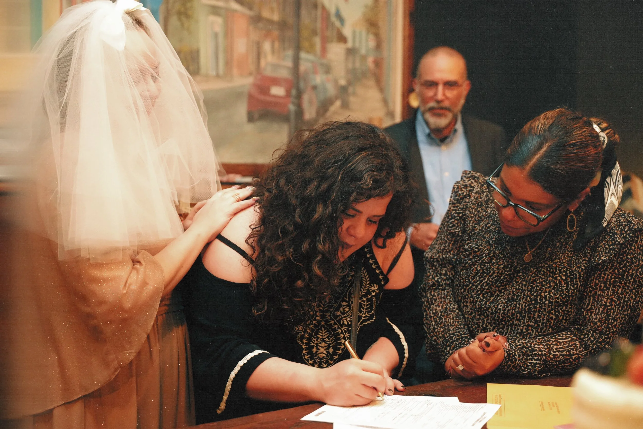 A woman in a black dress with embroidered details signs a document on a table as three women and a man observe. One of the women is dressed as a bride with a veil and gown, laying hands on her shoulder. A painting of a street scene hangs on the wall 