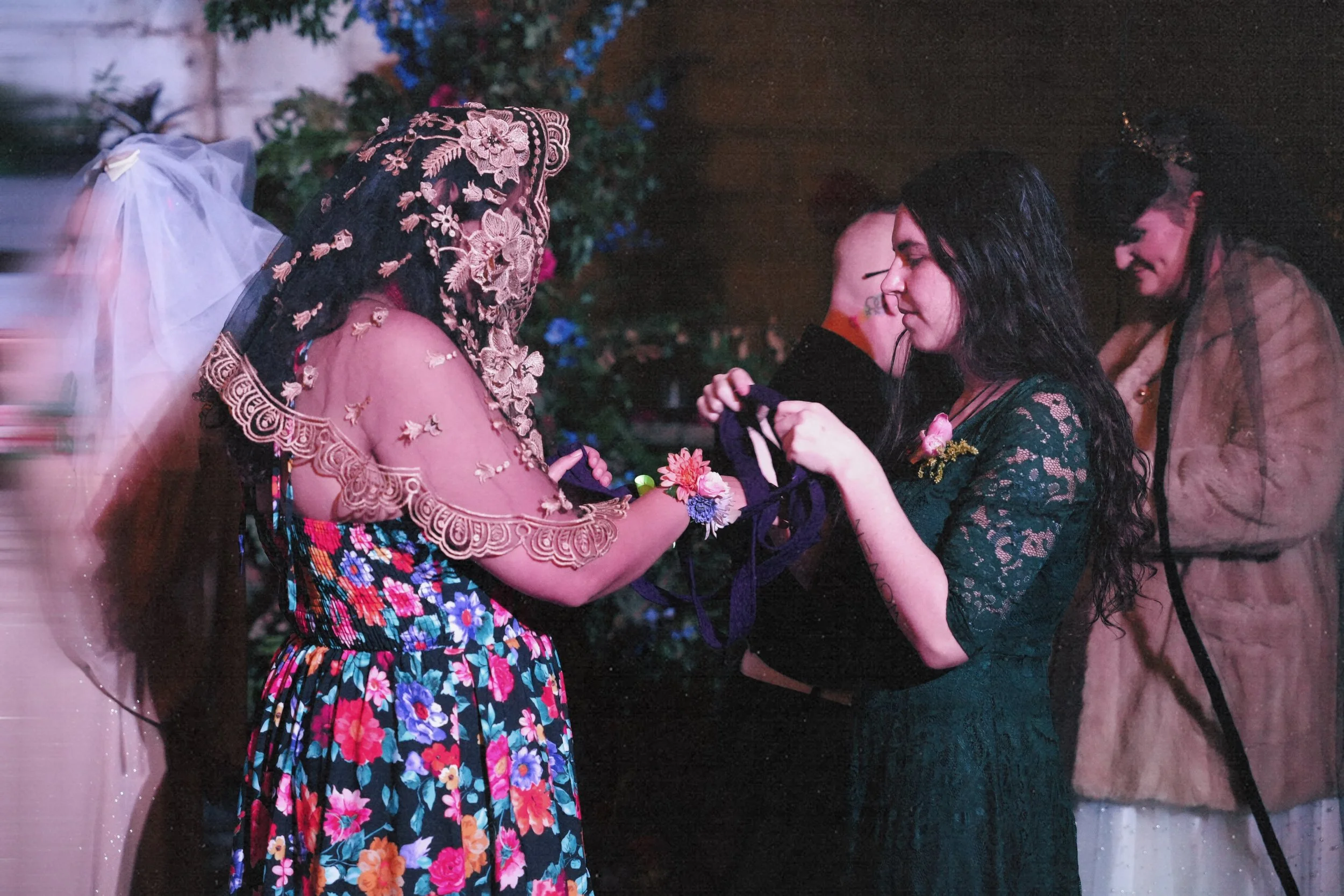 Group of women at a social event, one wearing a floral dress and lace shawl, receiving flowers or corsages from another woman in a green lace dress.
