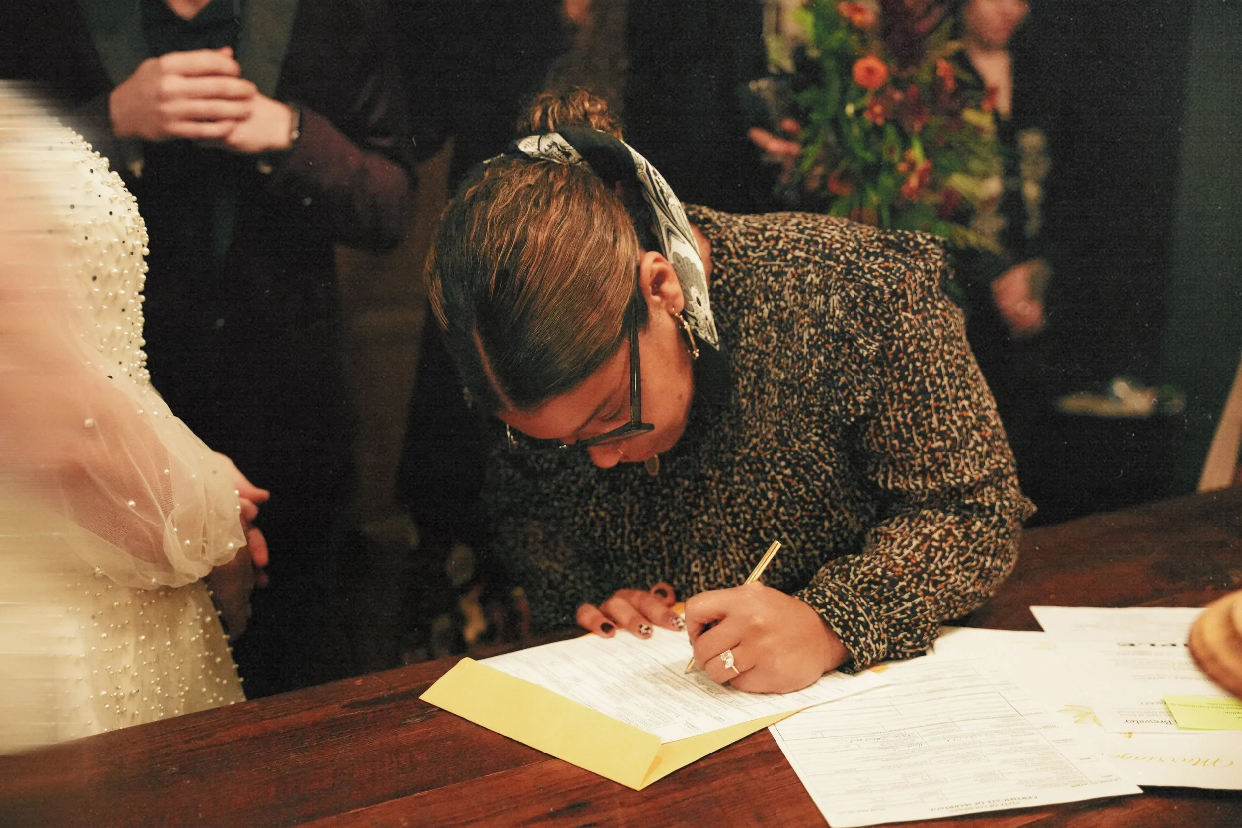 A woman signing a document at a table with a yellow paper, wearing glasses, a patterned top, earrings, and a headscarf, while others stand in the background.
