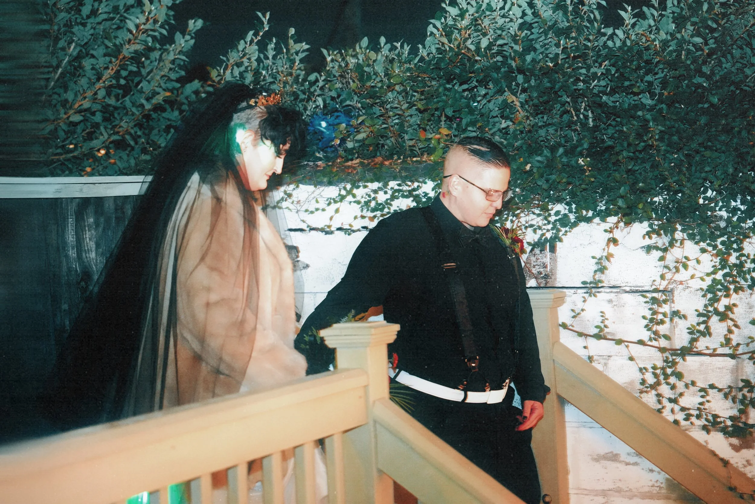 A woman dressed as a bride and a man dressed in black are sitting on a staircase outdoors. The woman is wearing a veil and the man has glasses and a distinctive hairdo. They appear to be in a contemplative moment with lush greenery in the background.