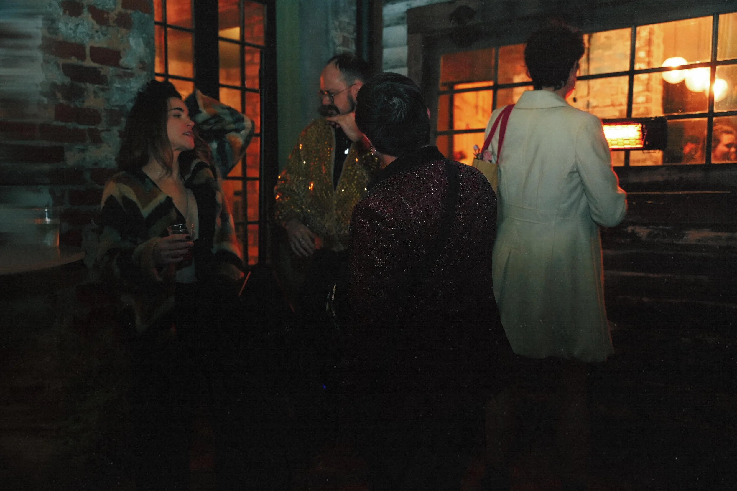 A group of four people socializing inside a dimly lit bar with brick walls and large windows, one woman holding a drink, two men and another woman engaged in conversation.