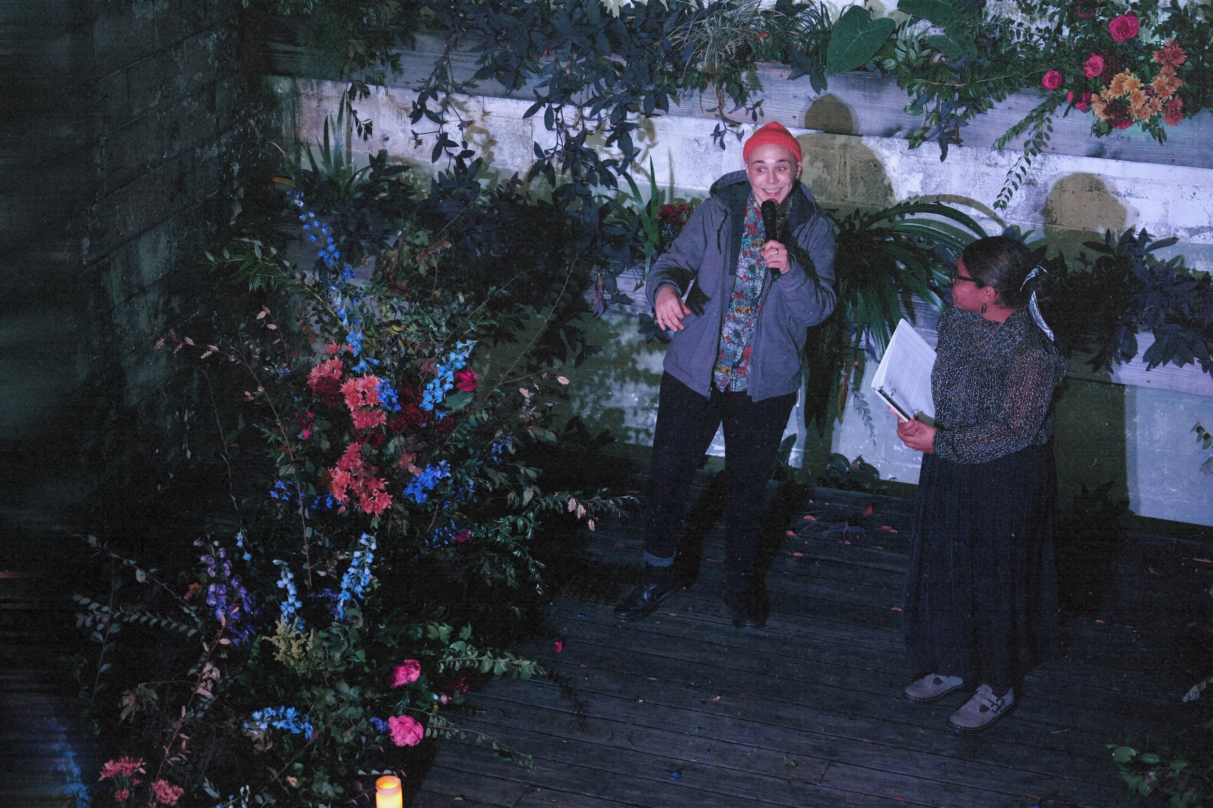 A woman with pink hair and a gray jacket speaking into a microphone next to another woman holding a notebook, outdoors with flowers and greenery.