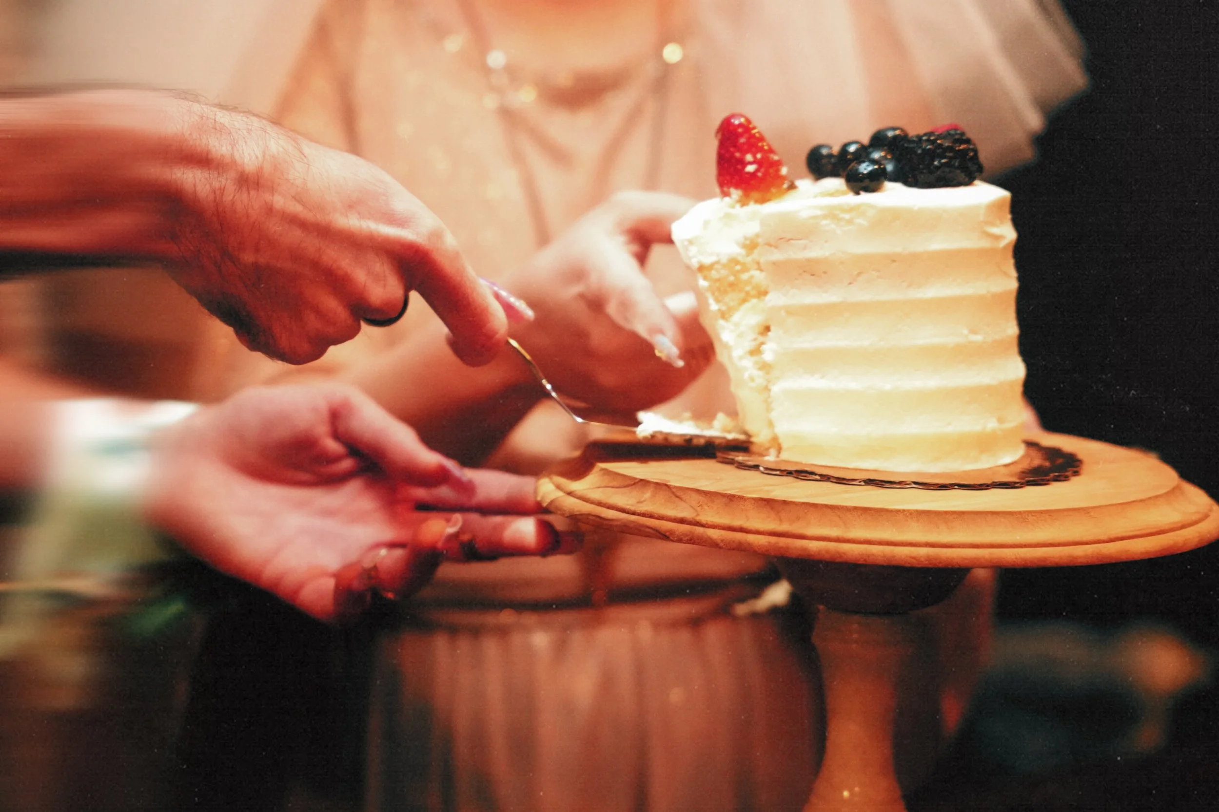 Close-up of hands slicing a layered white cake with cream frosting and black and red berries on top, on a wooden cake stand.