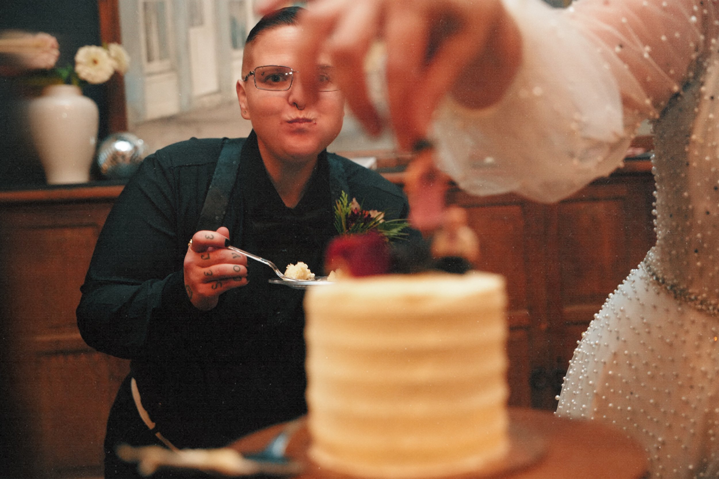 A person with glasses and tattoos holding a plate of cake in front of a woman in a beaded dress who is cutting a layered cake topped with berries and fruit, in a warmly decorated room.