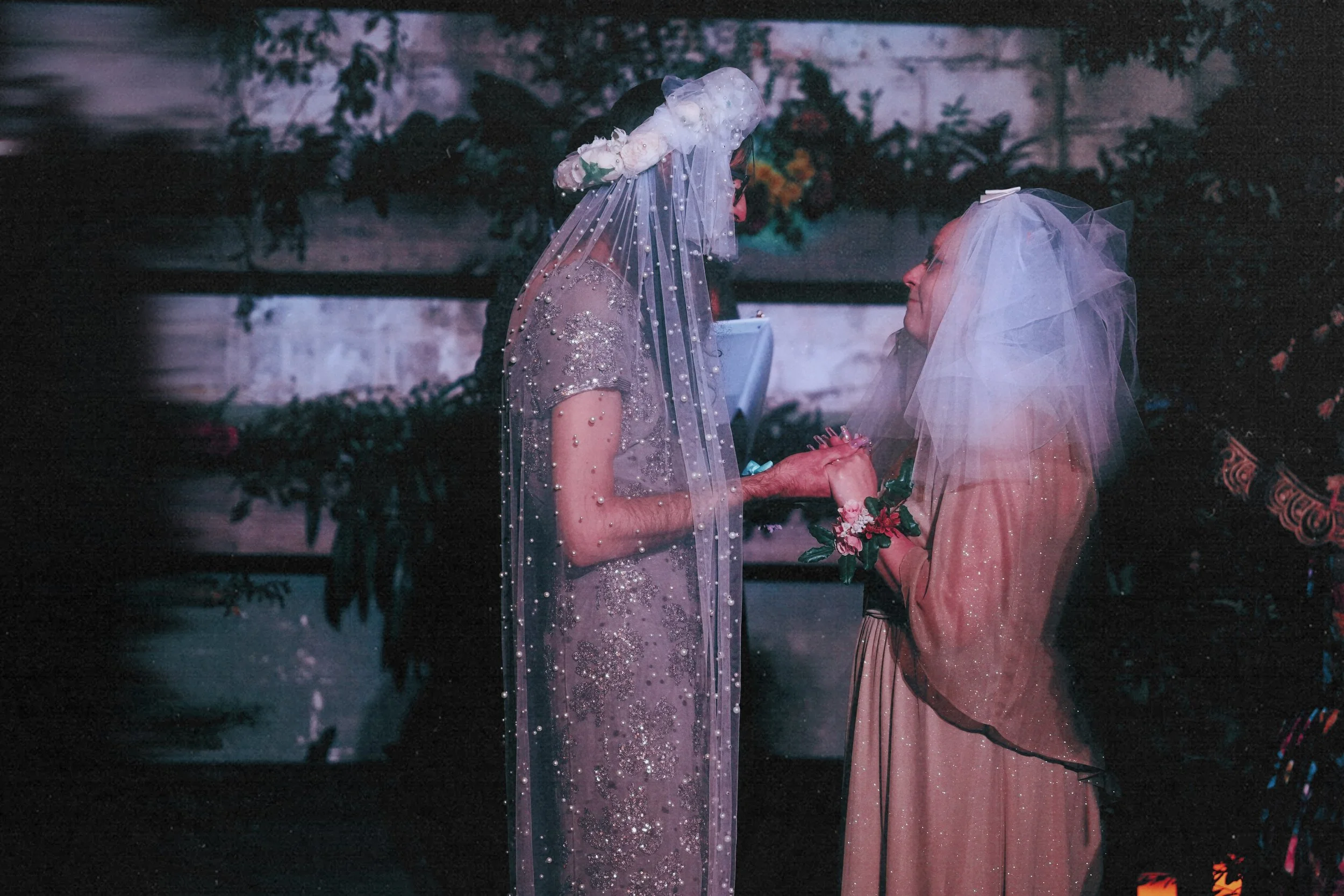 Two women dressed in vintage-style gowns and veils, holding hands and looking at each other, in a garden setting.