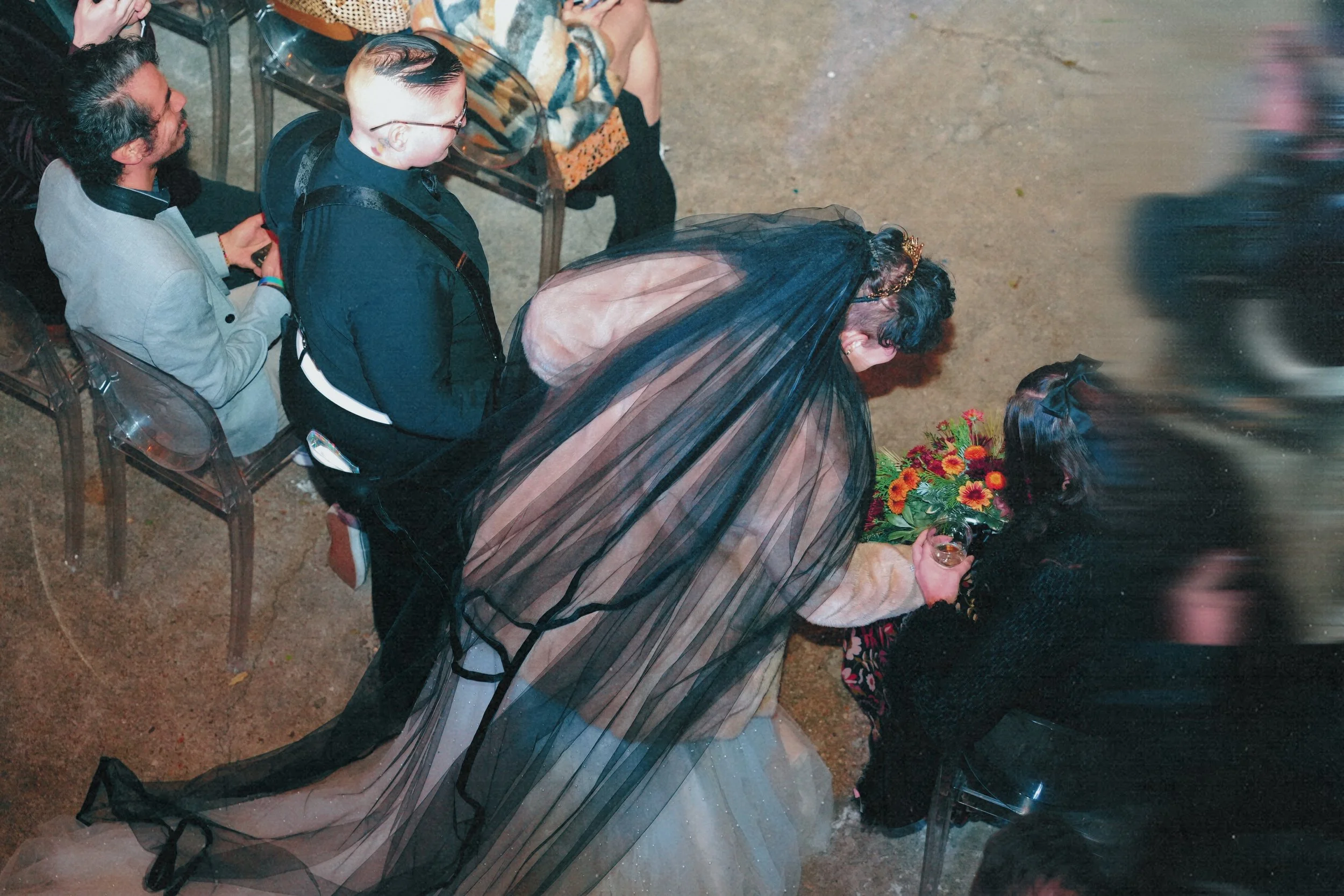 A bride with a long veil and a beige wedding dress is performing a handstand in front of a seated guest holding a bouquet of sunflowers, while other guests watch at a celebration or wedding reception.