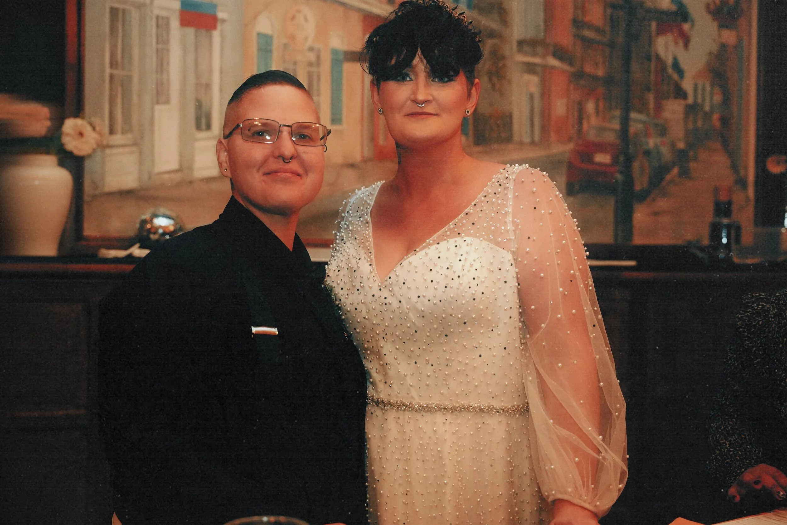 Two people standing together in a warmly lit room with a cityscape painting in the background. One is wearing a black outfit with glasses, and the other is in a white dress with pearl embellishments and sheer sleeves, both smiling at the camera.