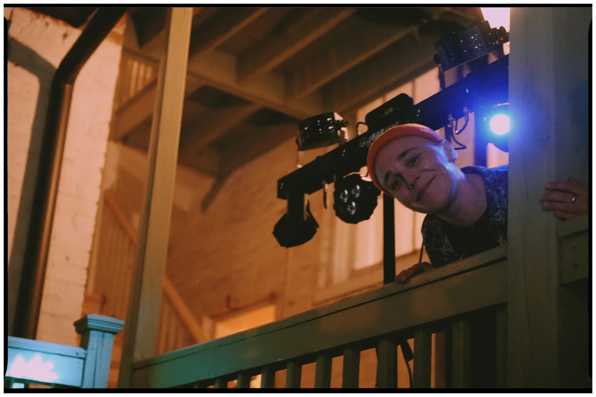 Person with glasses and orange beanie smiling while leaning over a balcony railing at a dimly lit indoor event.