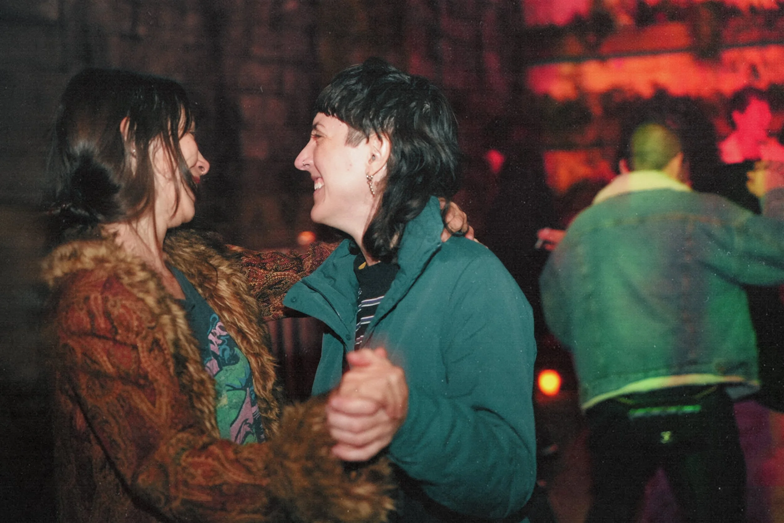 Two women are dancing close together in a dimly lit room, smiling at each other. One woman has dark hair and is wearing a furry coat, while the other woman has short dark hair and is wearing a teal jacket.