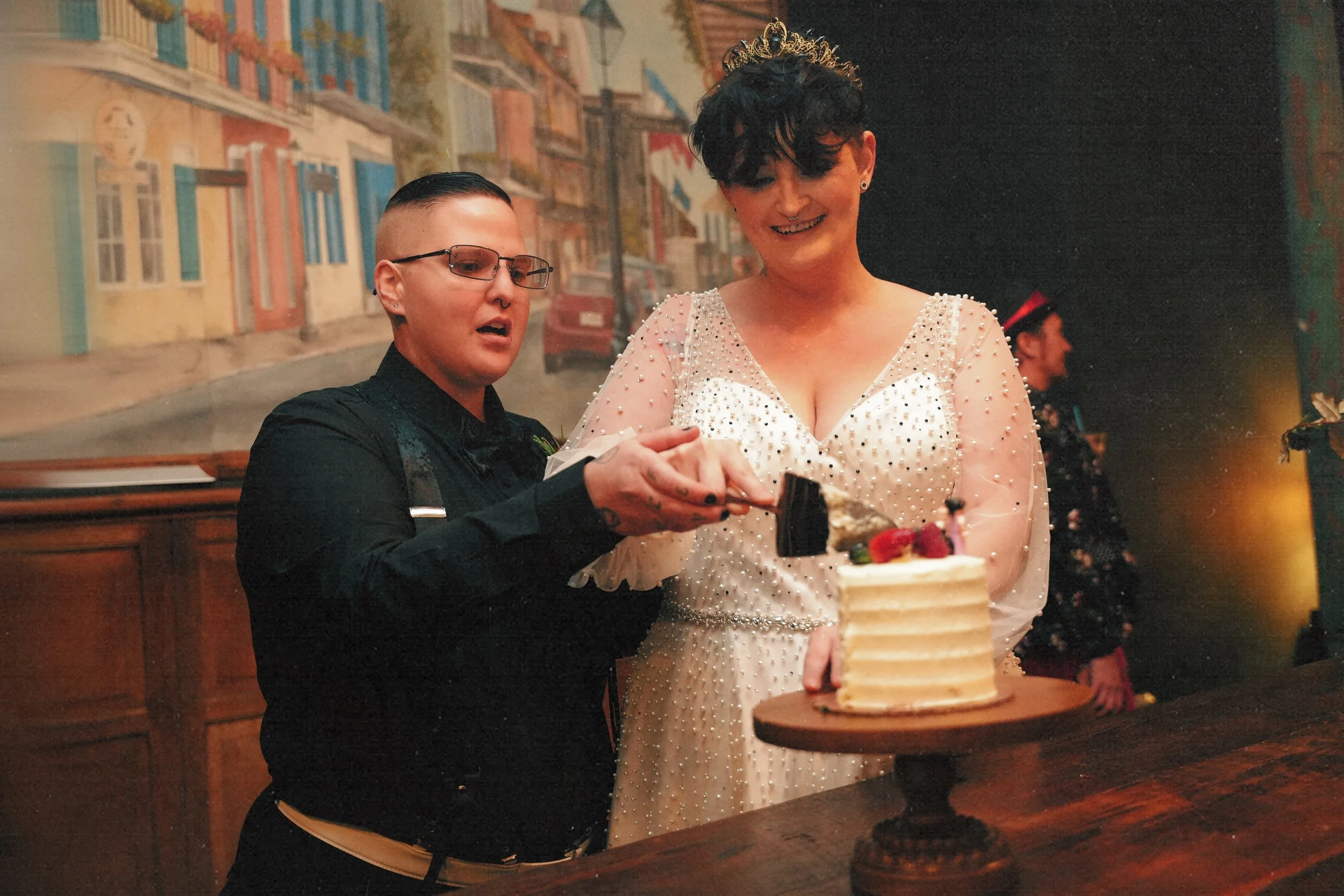 Two women, one with short hair and glasses and the other with short curly hair and a crown, are cutting a small white layered cake with strawberries on top. They are at a celebration, with a background of colorful buildings painted on a wall.