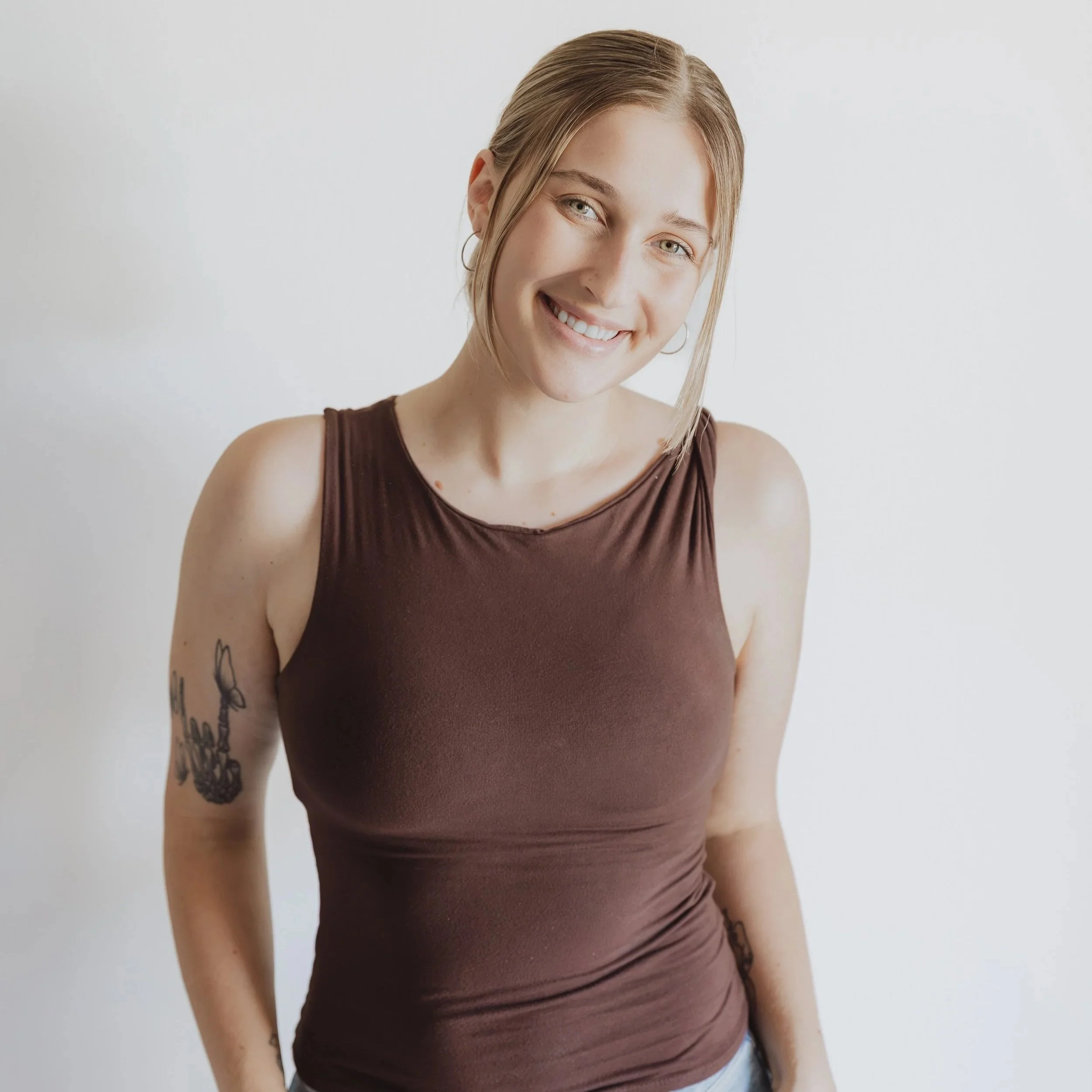 A smiling young woman with blonde hair and blue eyes wearing a sleeveless brown top standing against a plain white background.