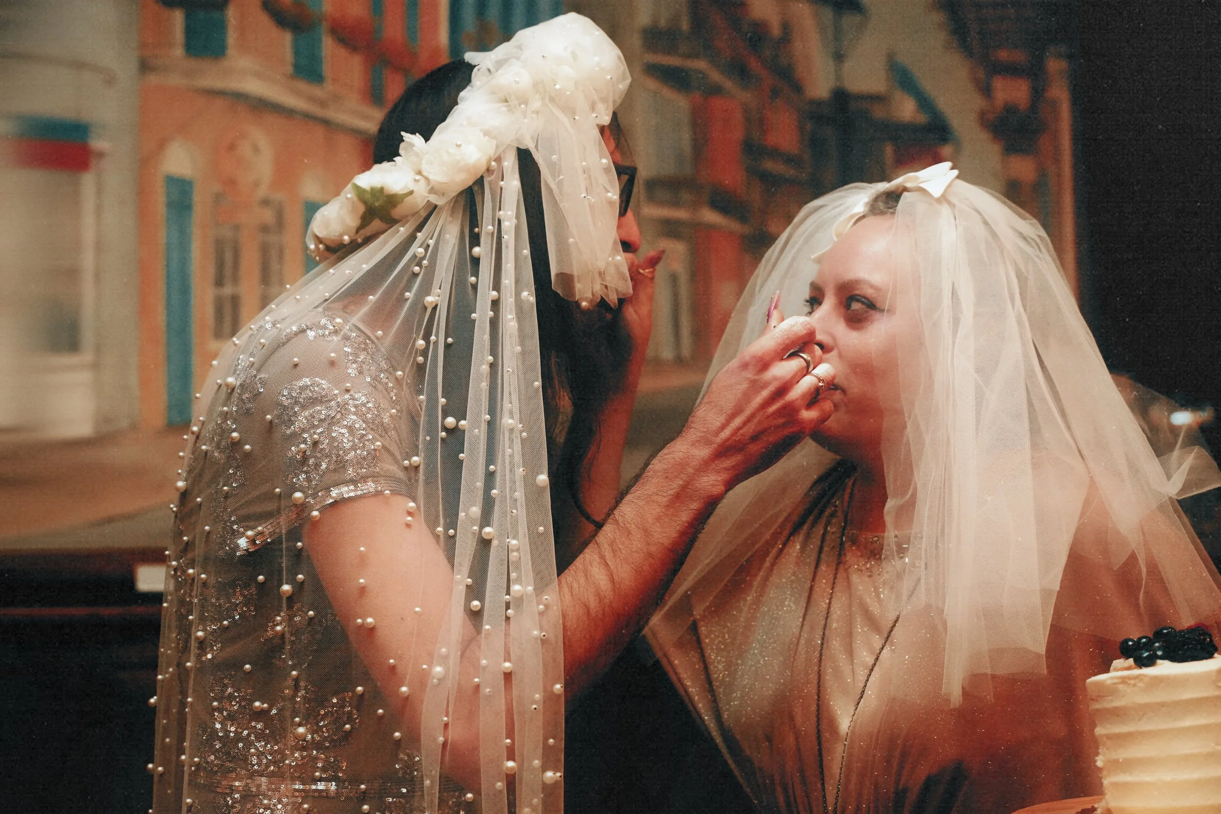 Two women, one with dark hair and glasses and the other with blonde hair, are at a celebration or wedding. The woman with dark hair is gently touching the face of the blonde woman who is wearing a veil. There is a cake with berries visible in the for