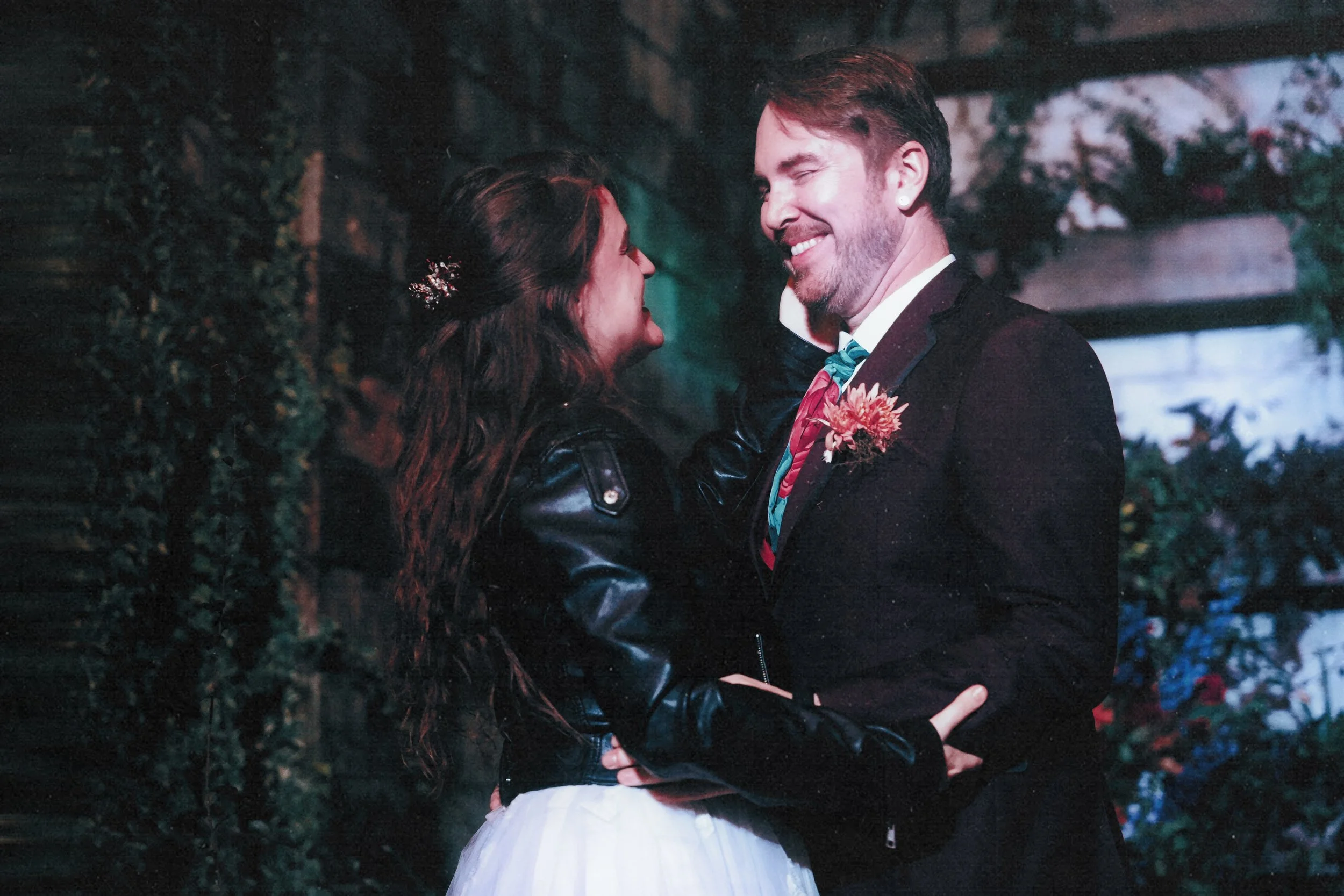 A man and woman are smiling and facing each other, in an outdoor setting with greenery and flowers in the background. The woman has long, curly hair and is wearing a leather jacket and a white dress. The man has short hair and is dressed in a dark su