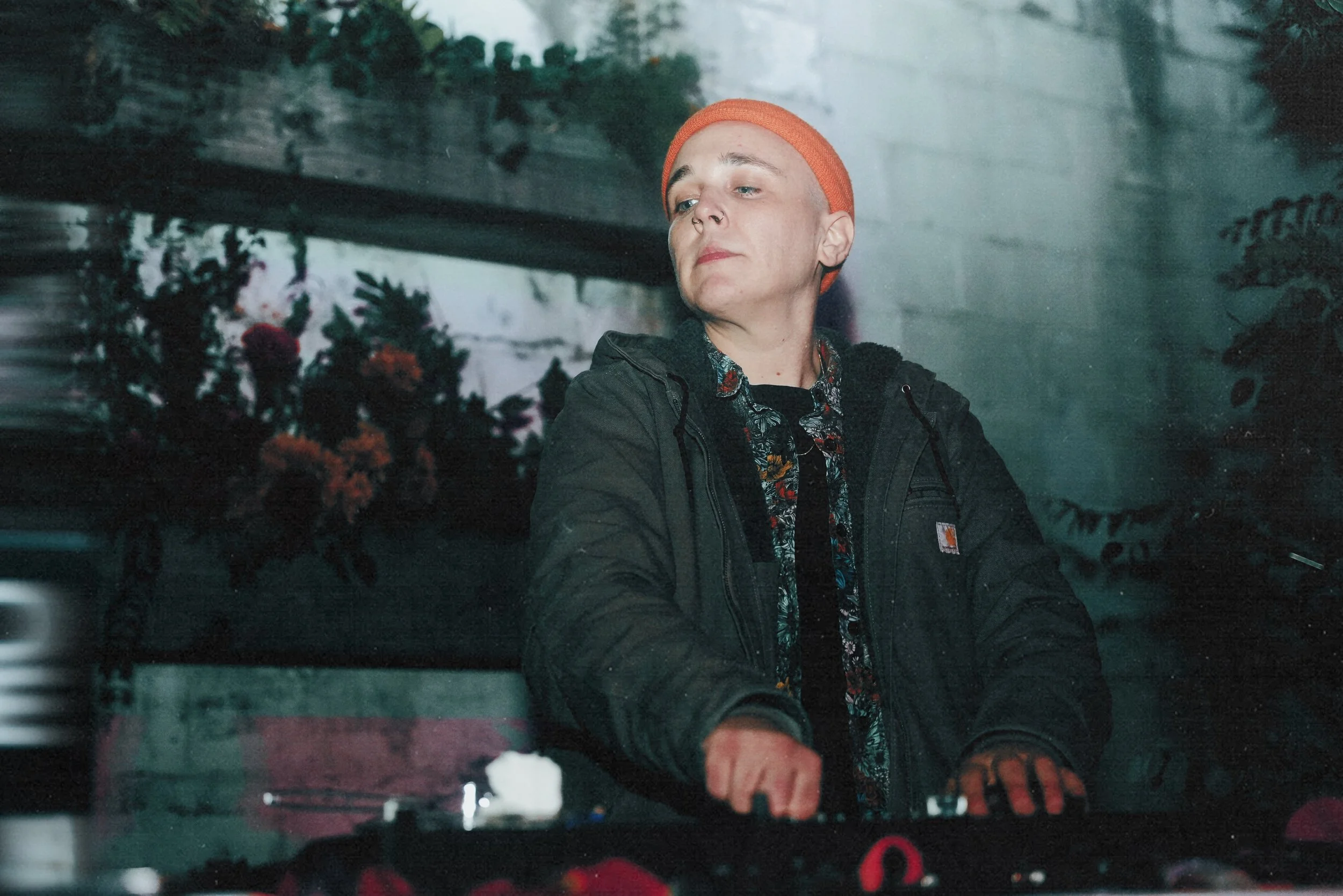 A person with a shaved head wearing an orange beanie, black jacket, and vibrant shirt, appears to be DJing in a dimly lit setting with plants in the background.