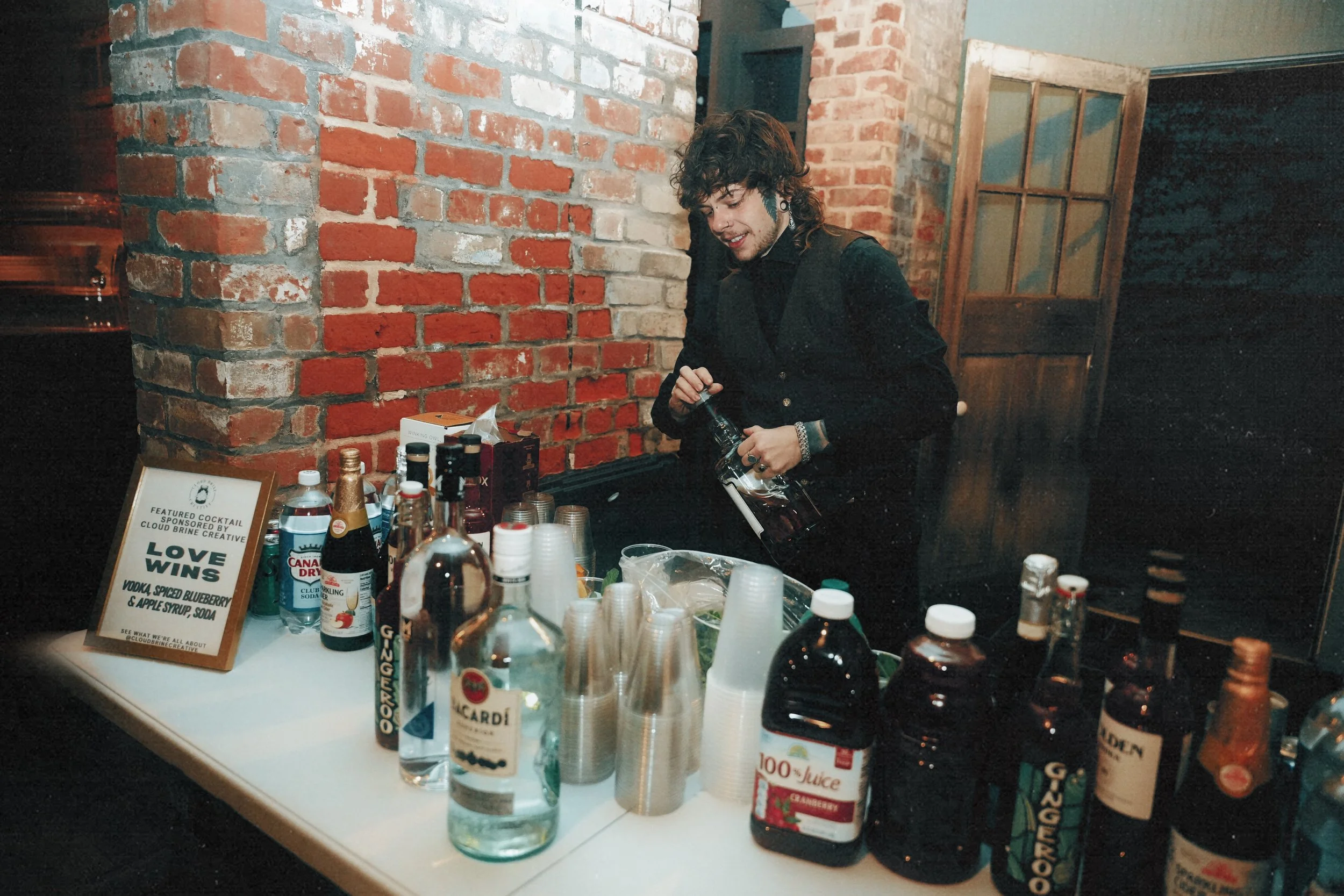A woman preparing a cocktail at a bar with brick walls. The table has various bottles of alcohol, juices, and cups. A sign on the table advertises a cocktail called 'Love Wins' with vodka, blueberry syrup, and apple syrup.