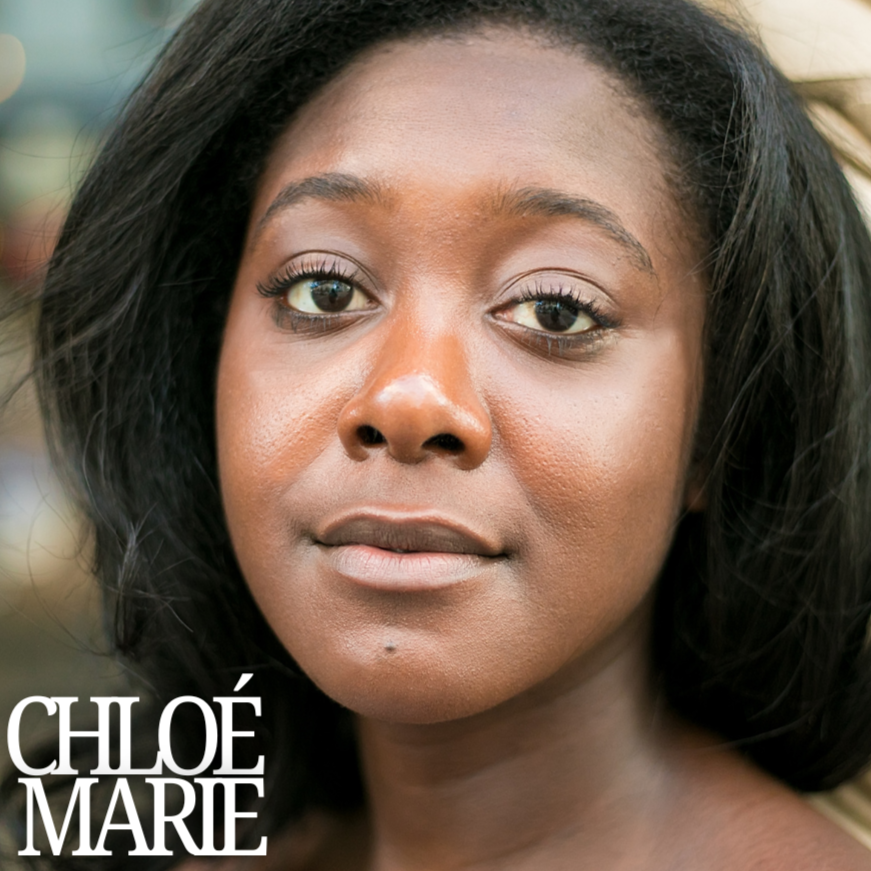 Close-up of a woman with medium brown skin, dark hair, and neutral expression, outdoors with blurred background.