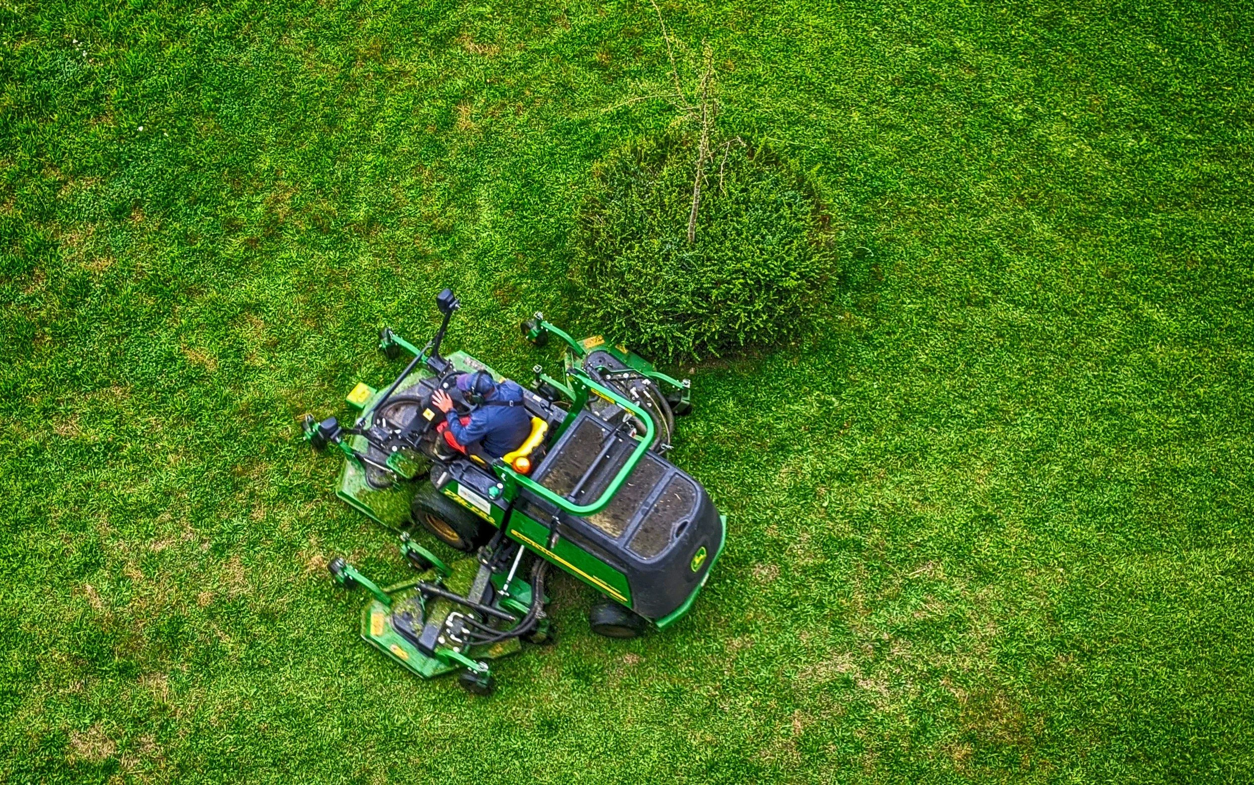 An aerial view of a person in a blue jacket operating a green riding lawn mower on a grassy area with a large shrub nearby.