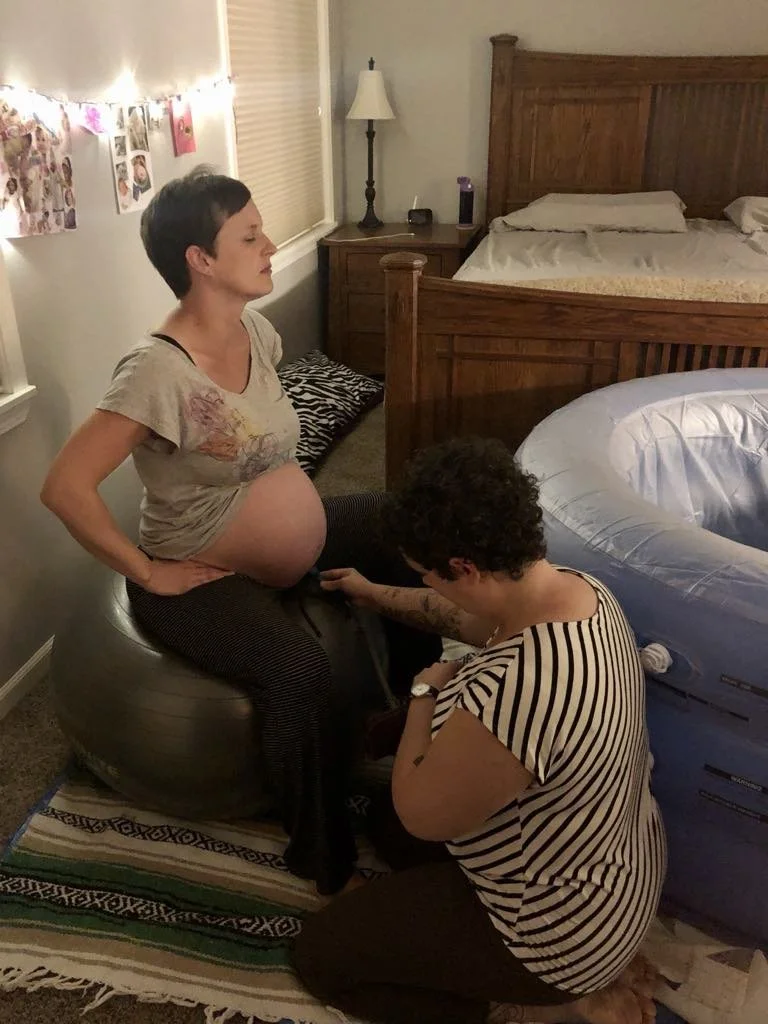Pregnant woman sitting in her bedroom on a gray exercise ball next to a birth tub while midwife Noel Fernandez kneels in front of her, listening to her baby's heartbeat with a doppler.