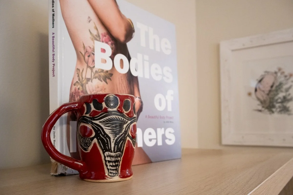 Decorative red mug with a black and white uterus design, resting on a wooden surface beside a book titled 'The Bodies of Mothers' and a framed floral print.