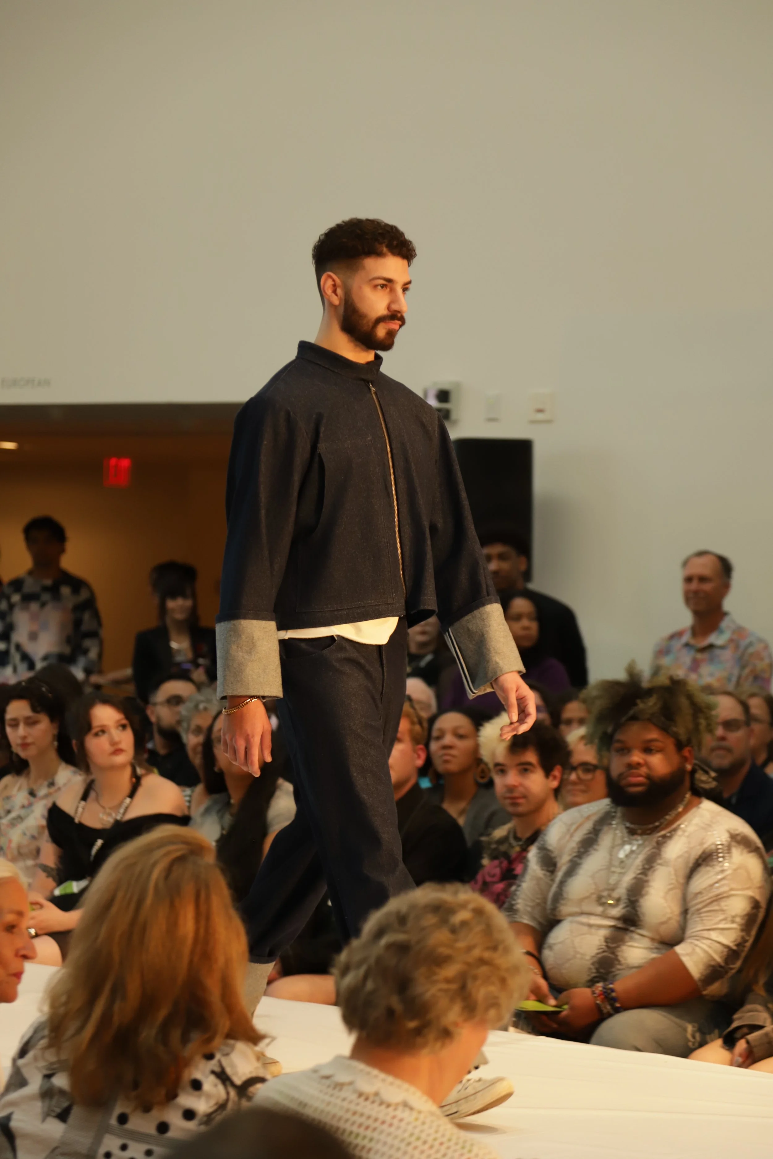 Model walking on a runway, wearing a dark outfit with rolled-up sleeves, in front of an audience.