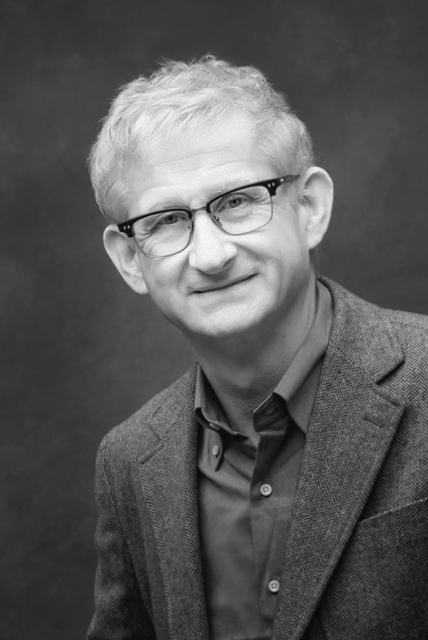 Black and white portrait of a middle-aged man with glasses, light hair, wearing a blazer and collared shirt, smiling.