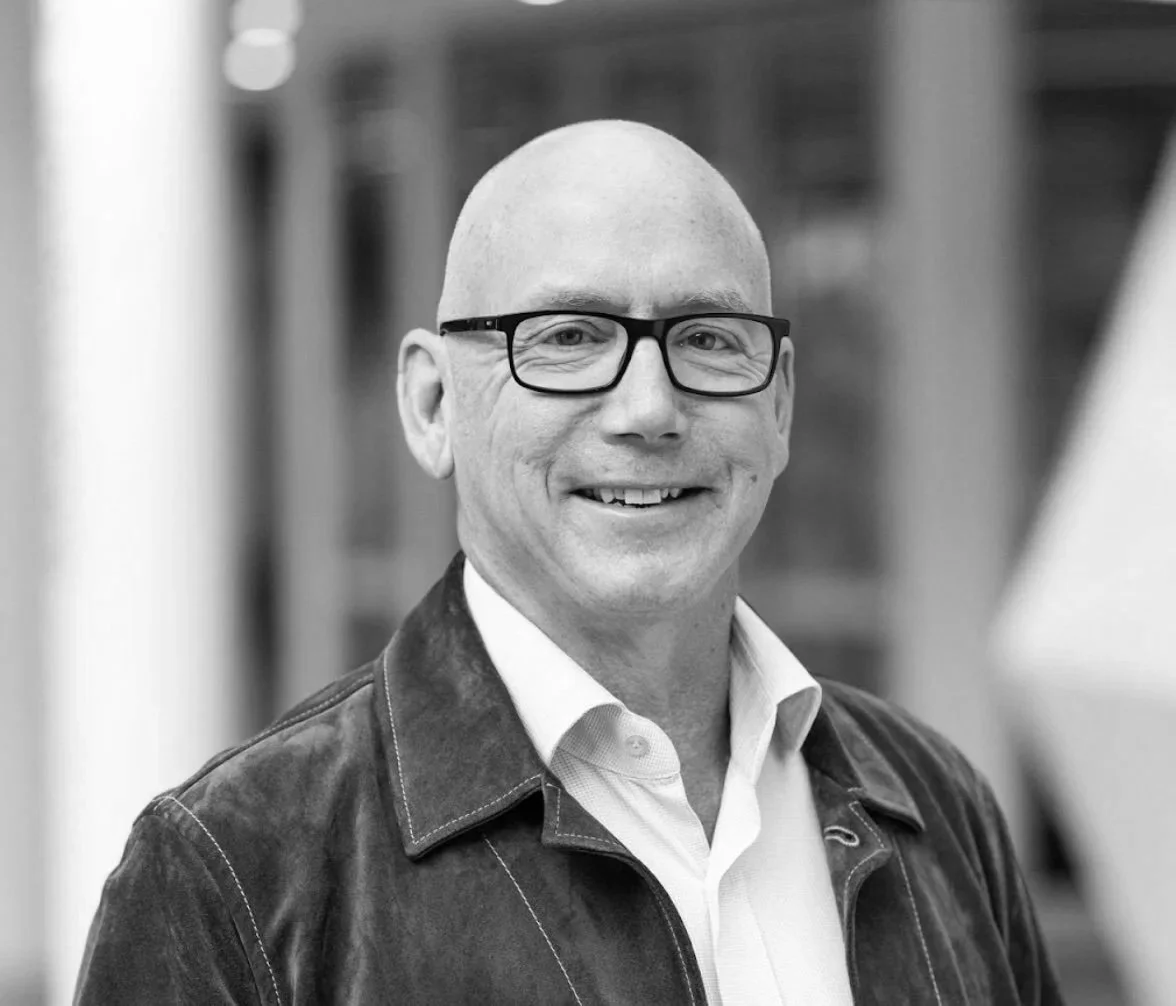 Black and white photo of a smiling bald man wearing glasses, a white collared shirt, and a dark jacket, standing outdoors with a blurred background.