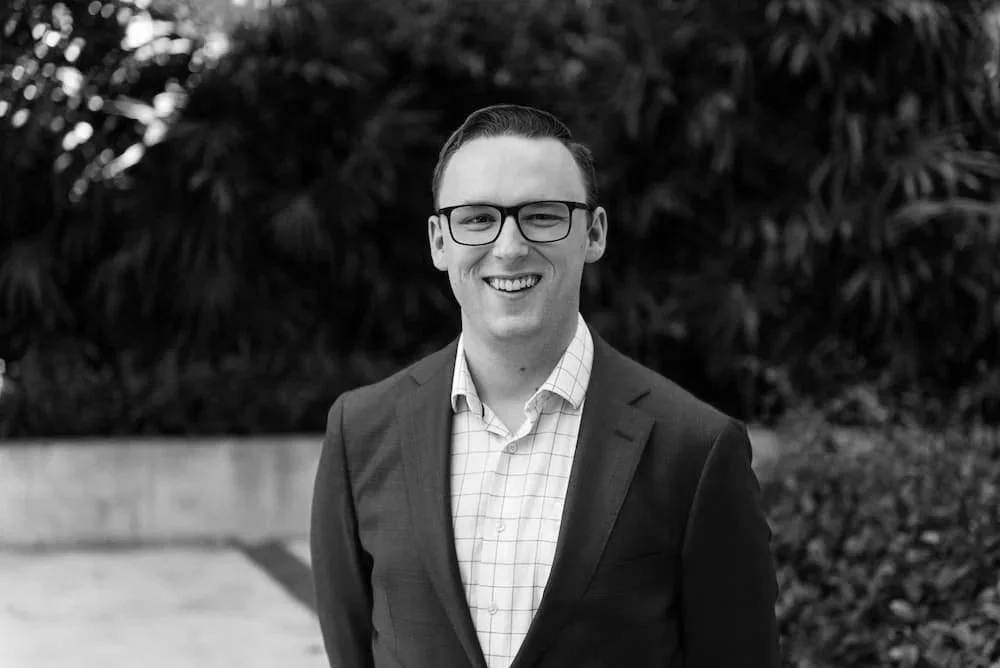 A smiling man with glasses, wearing a suit and a checkered shirt, standing outdoors with trees in the background.