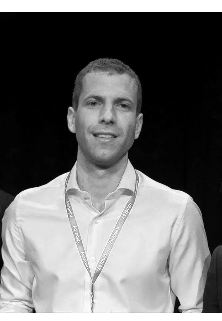 A young man in a light-colored dress shirt wearing a lanyard with 'Mindray' written on it, smiling at the camera.
