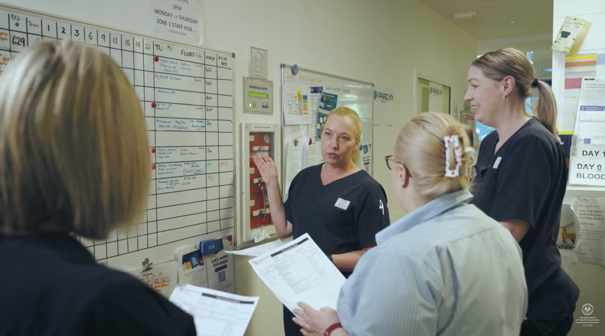 Healthcare professionals and patients gathered in a hospital corridor near a whiteboard with schedules and notes, engaging in a discussion.