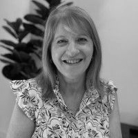 Black and white photo of a smiling woman with shoulder-length hair, wearing a floral patterned blouse, standing in front of a leafy plant.